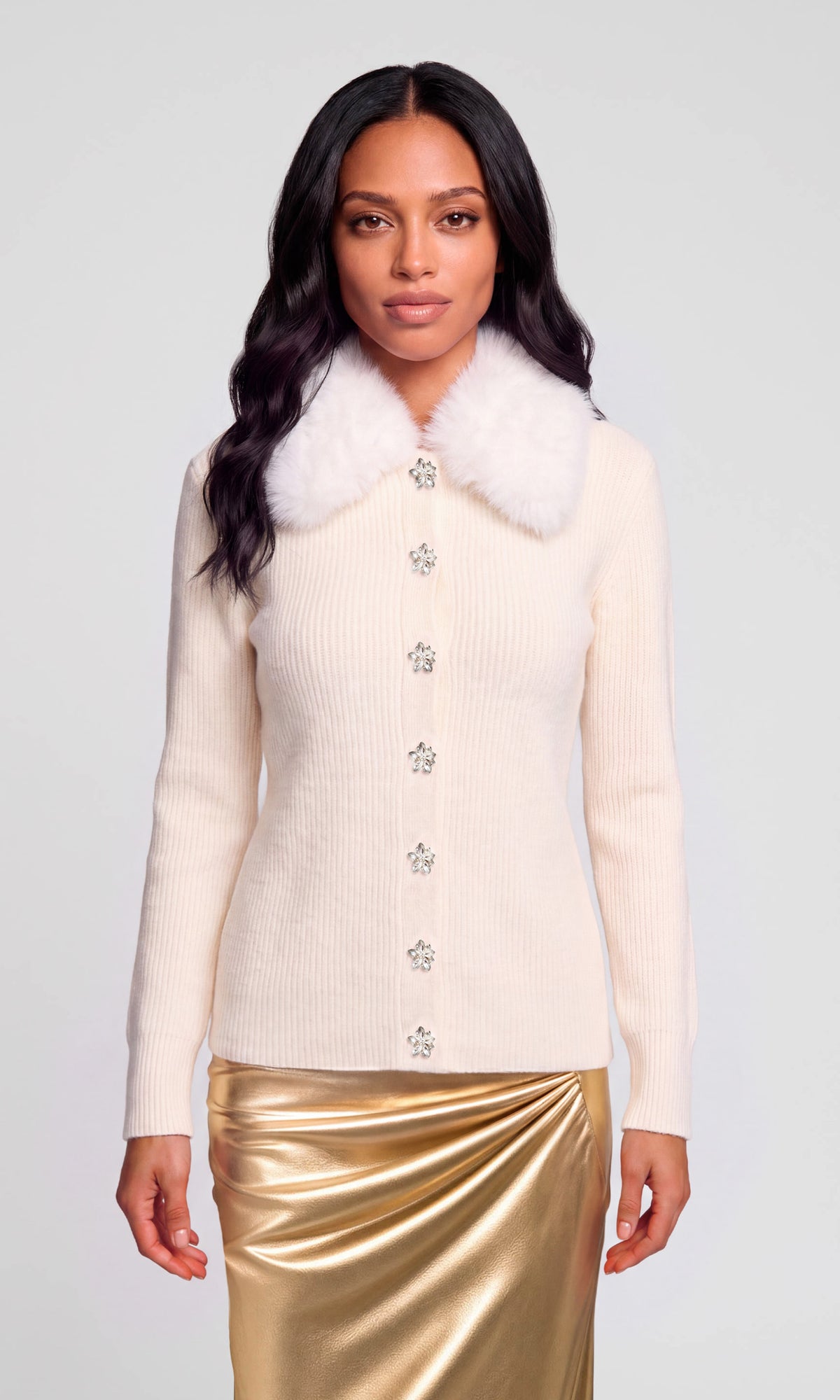 Model wearing a cream ribbed knit cardigan with a fluffy white faux-fur collar and decorative crystal-like buttons, paired with a metallic gold skirt.