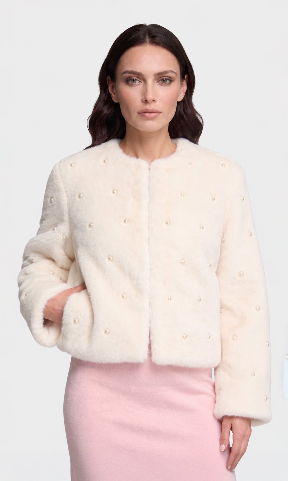 A woman modeling a cropped, cream-colored faux fur jacket with small pearl-like bead details, paired with a light pink dress.