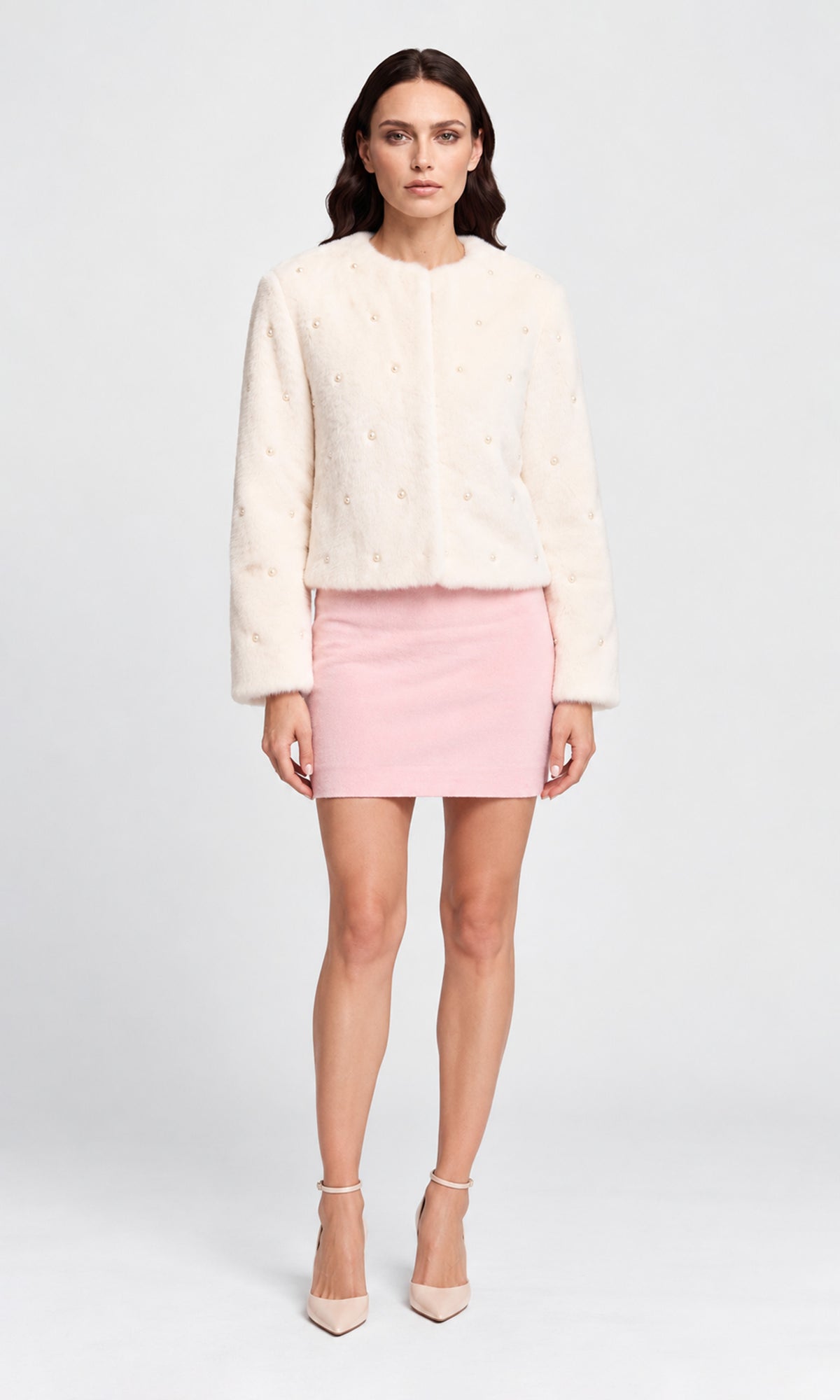 Model wearing a cream faux-fur cropped jacket with subtle pearl embroidery, a light pink mini skirt, and nude high heels.