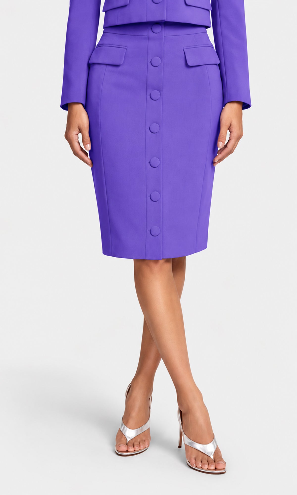 A model wearing a vibrant purple knee-length pencil skirt with a button-front closure and flap pockets, paired with a matching purple jacket and white heeled sandals.