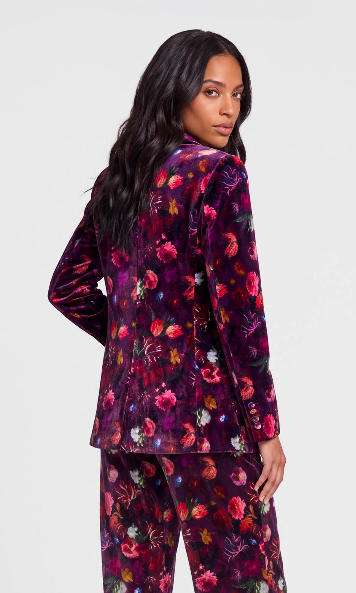 Back view of model wearing a dark floral velvet blazer and matching trousers against a light gray studio background.