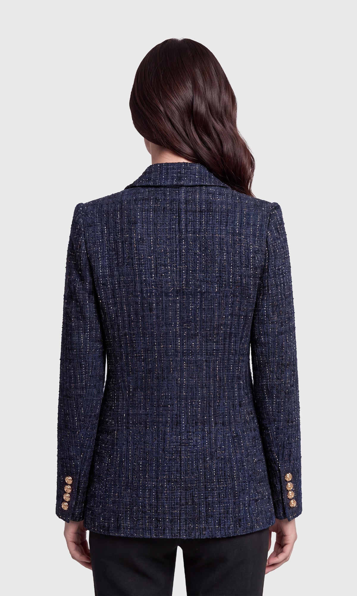A woman viewed from the back wearing a navy blue tweed blazer with gold buttons on the cuffs, paired with black pants.