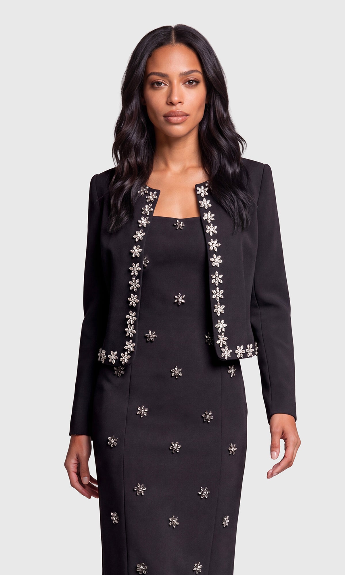 A woman modeling a black tailored jacket and matching long dress set, both decorated with silver-toned floral applique.