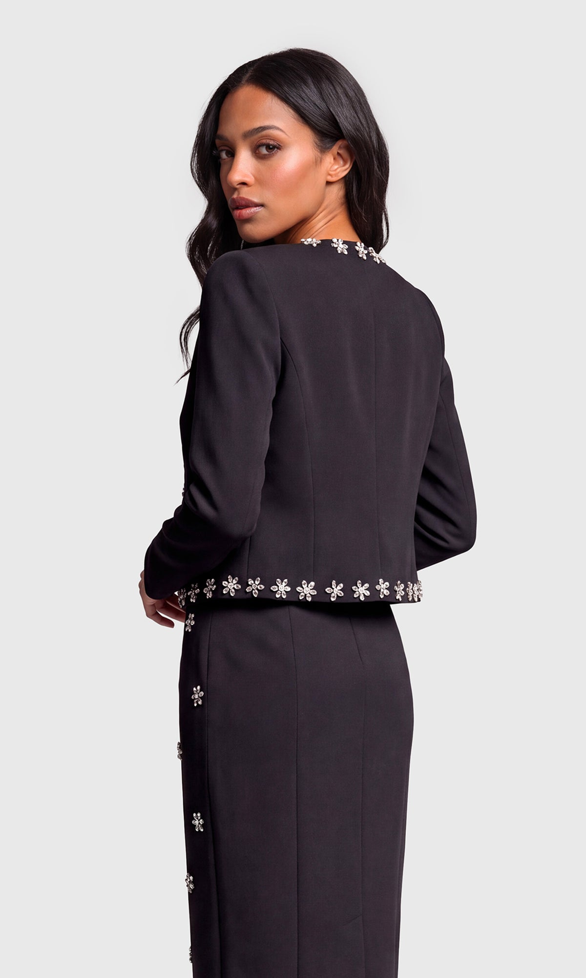 A woman shown from the back wearing a black long-sleeve dress with decorative silver floral embellishments along the neckline, waist, and side seams.