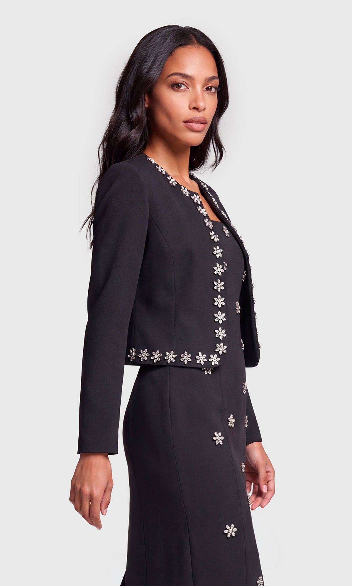 A woman modeling a black long-sleeve jacket and matching skirt set with white embroidered floral details.