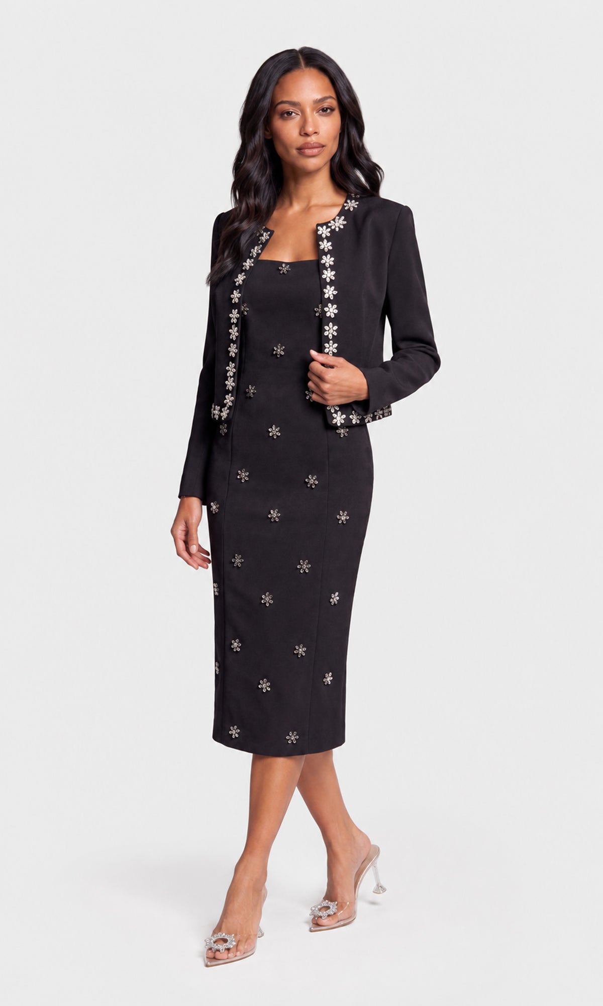 A woman models a black two-piece suit set consisting of a knee-length sheath dress and a matching long-sleeve blazer, both decorated with silver floral embroidery.