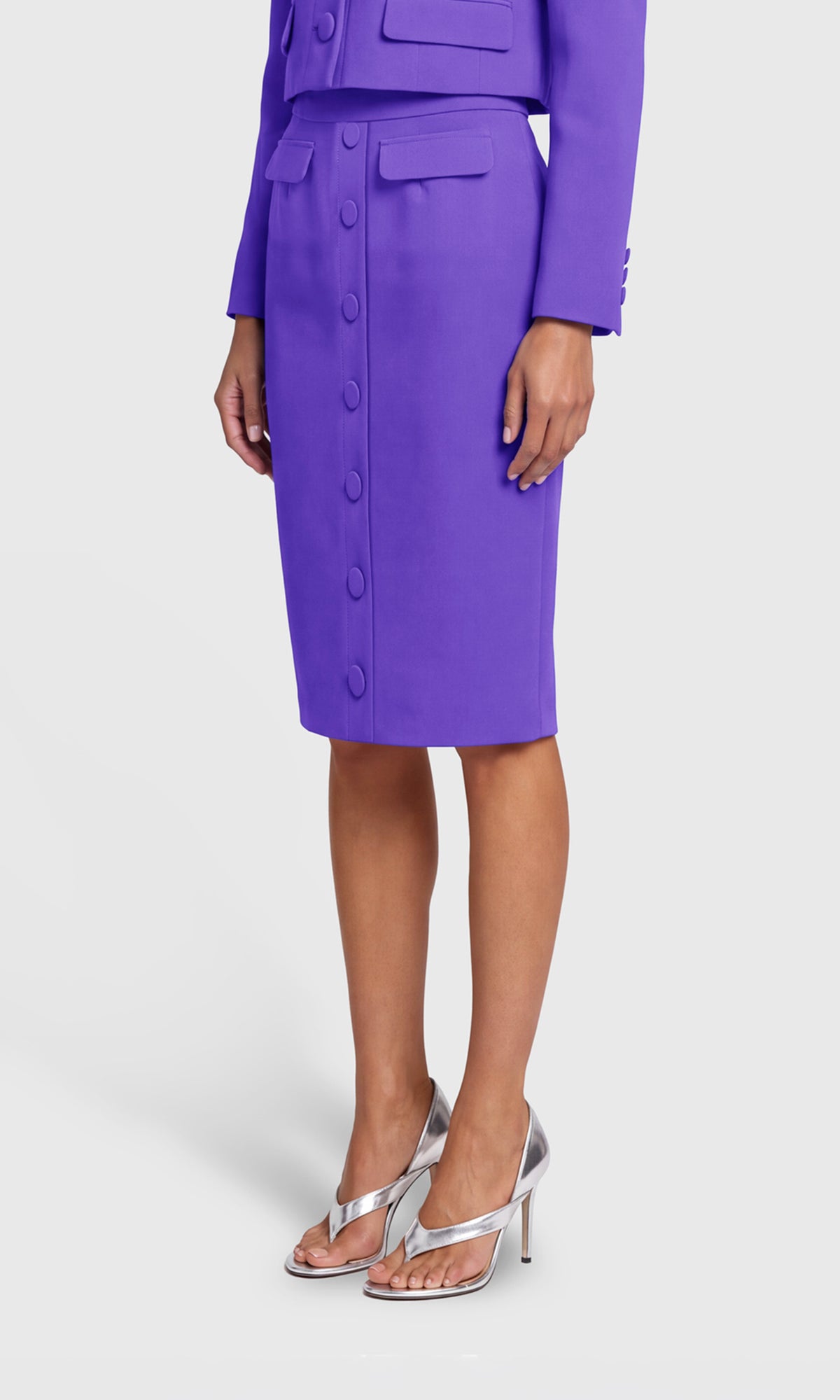 A model wearing a vibrant purple knee-length pencil skirt with a button-front detail, paired with a matching purple blazer and silver thong sandals.