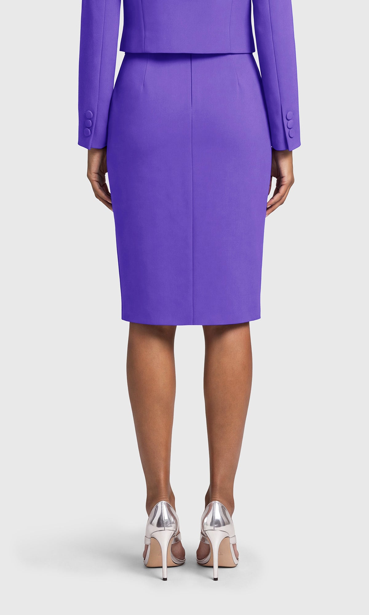 A model shown from the back wearing a vibrant purple knee-length pencil skirt with a matching blazer and clear high-heeled sandals.