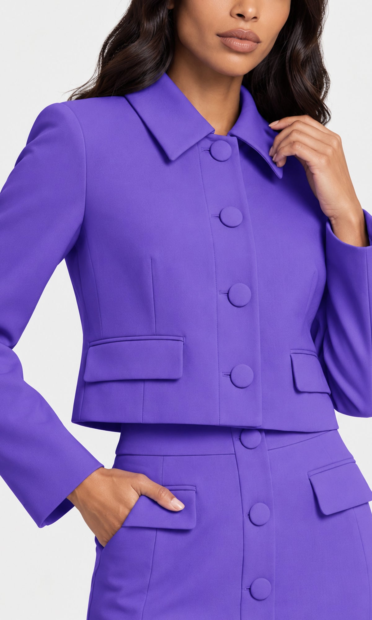 A close-up of a model wearing a vibrant purple tailored jacket and matching skirt set. The jacket features a structured collar, large decorative buttons down the front, flap pockets on the waist, and matching buttons on the front of the skirt.