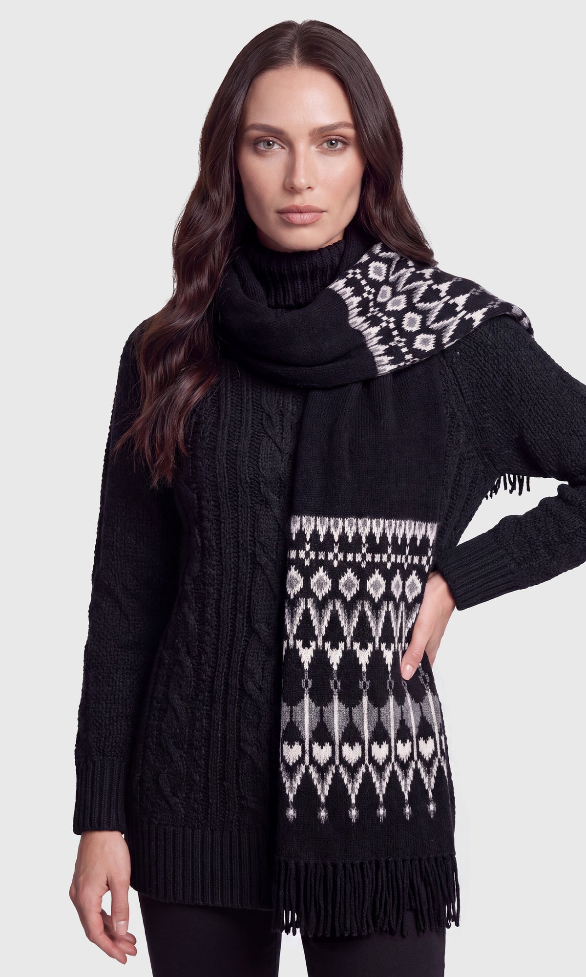 A woman modeling a black cable-knit turtleneck sweater with a matching black scarf featuring a white and gray geometric Fair Isle pattern and fringe details.