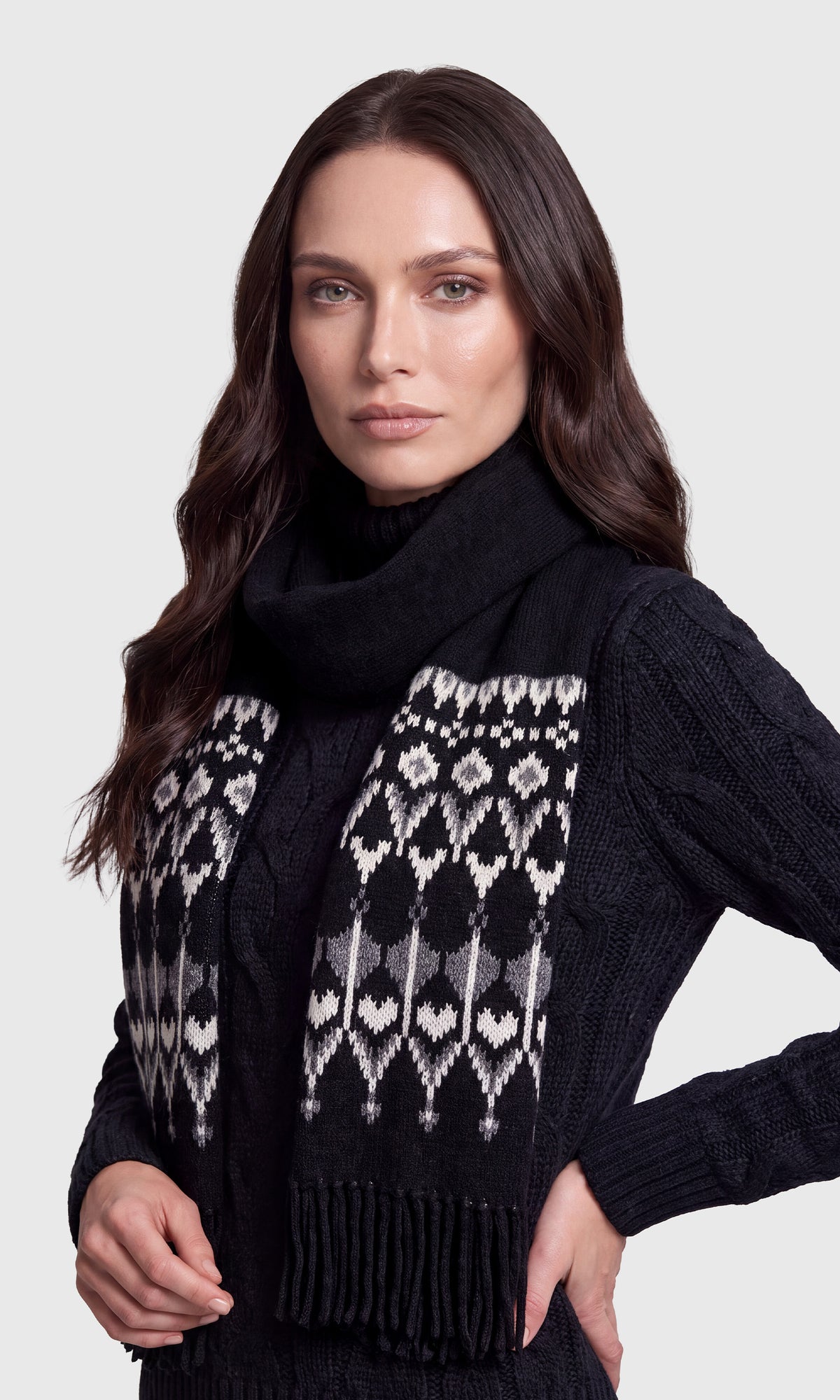 A woman wearing a black knit sweater and a black scarf with a white and gray geometric Fair Isle pattern and fringe.