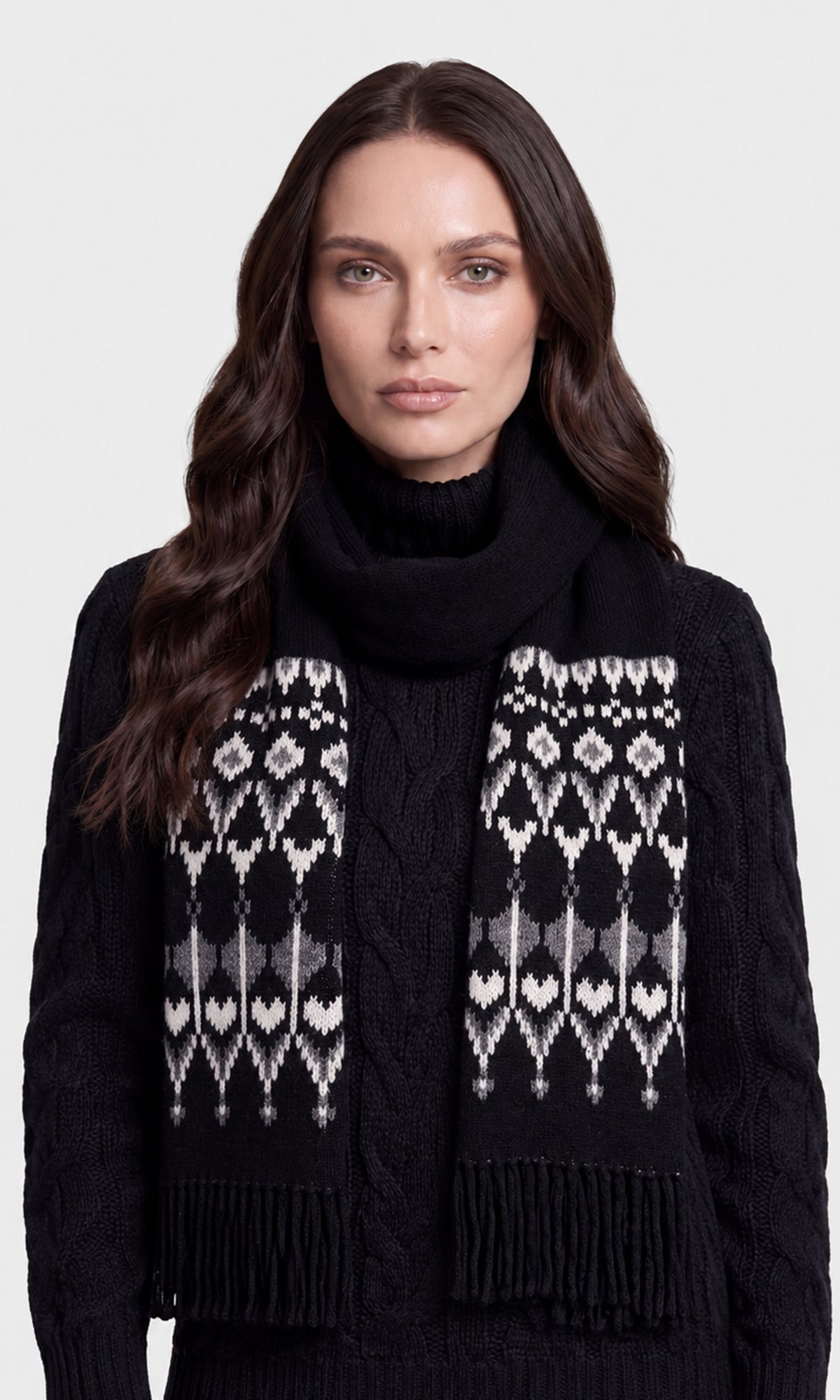 A woman wearing a black knit turtleneck sweater and a matching black scarf with cream and gray geometric patterns and fringe.