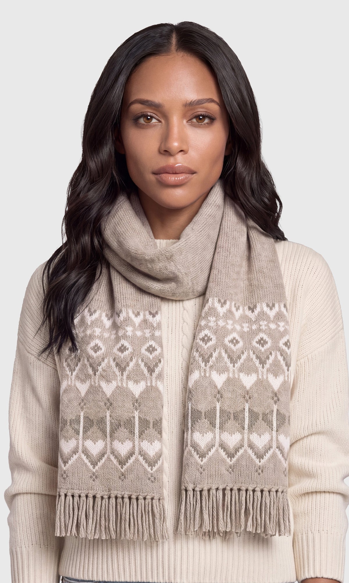 A woman wearing a beige and cream patterned knit scarf with fringe, styled over a cream ribbed sweater against a light gray studio background.