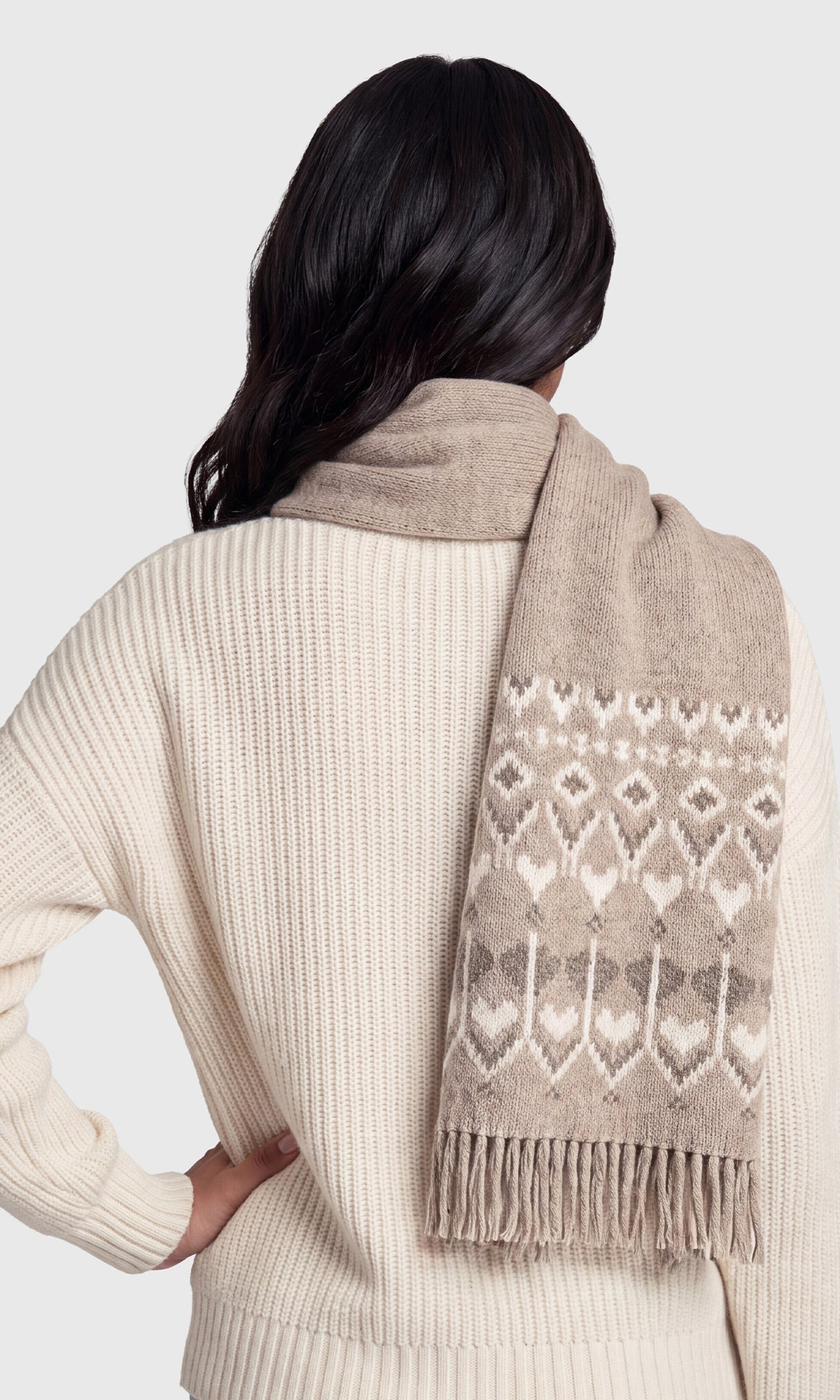 A woman shown from the back wearing a cream ribbed knit sweater and a beige scarf with a brown and cream geometric pattern and fringe.