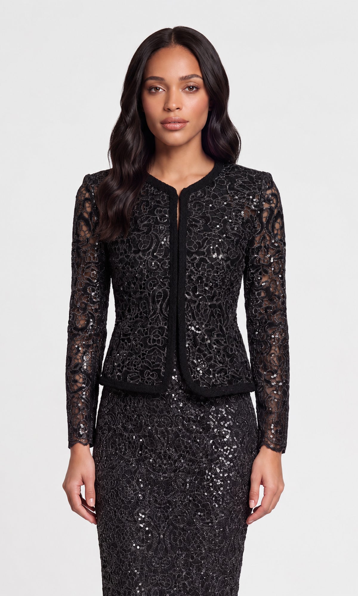 A woman modeling a black sequined lace jacket and matching skirt set against a light gray studio background.
