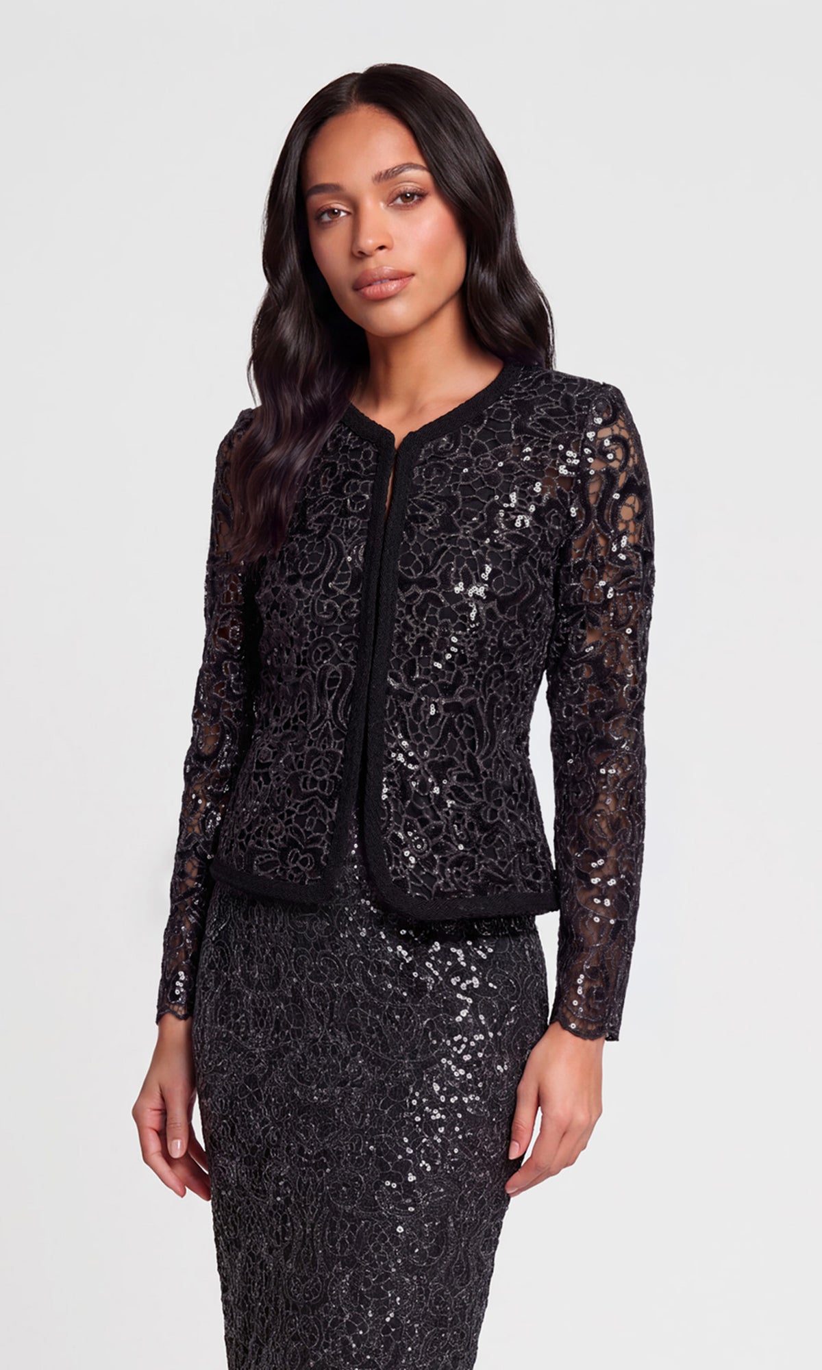 A woman modeling a black sequined lace jacket and matching skirt set against a light gray studio background.