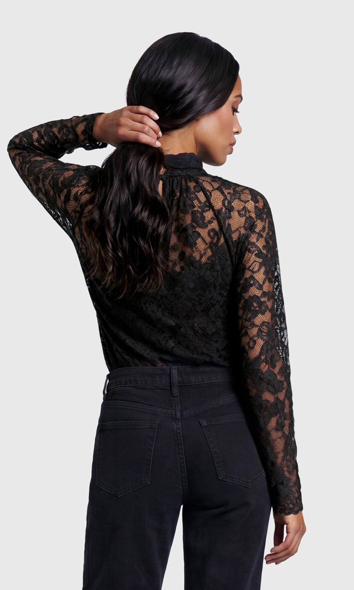 Back view of model wearing a black lace, mock-neck top with black jeans.