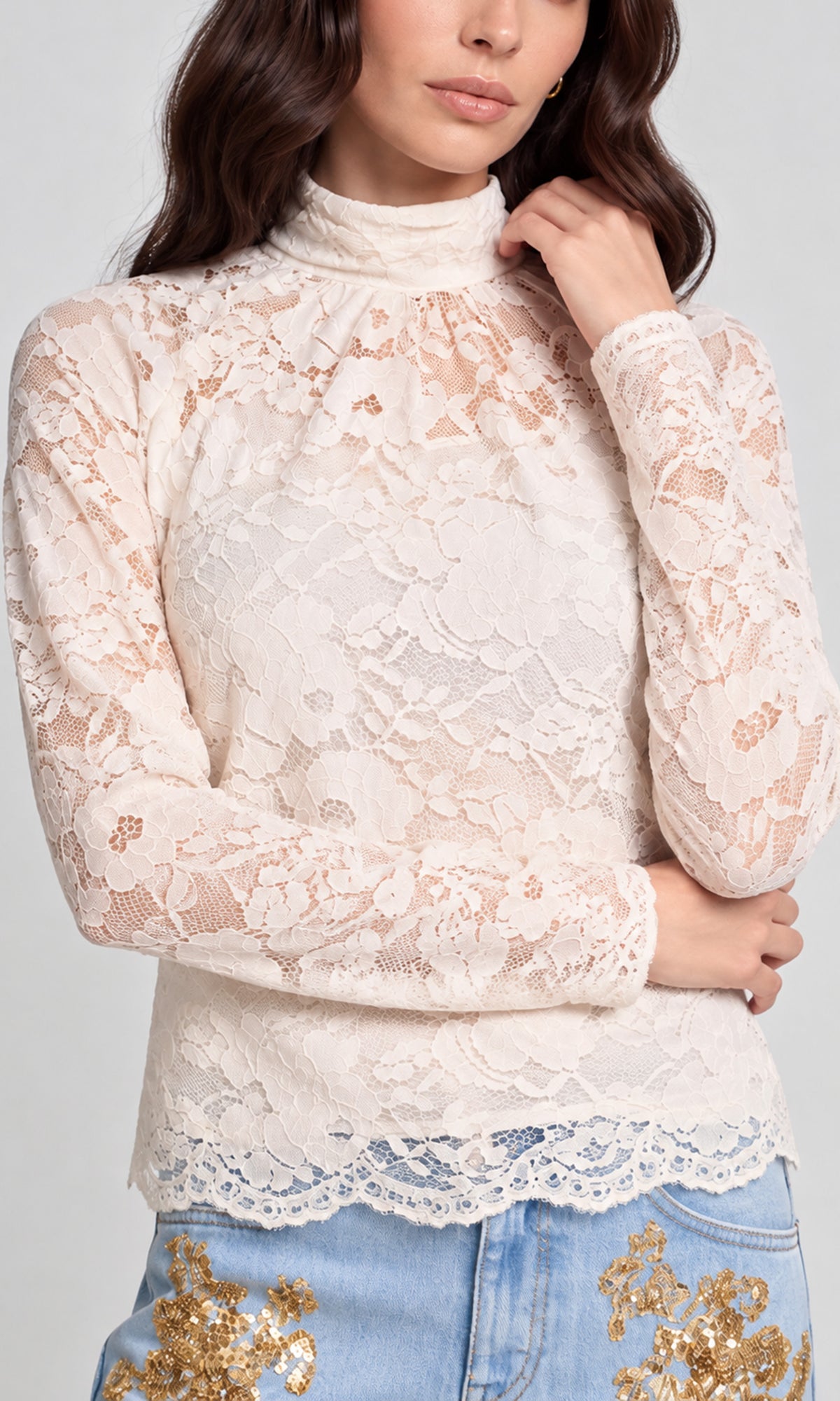 Close-up of  a cream lace mock neck long-sleeve top paired with light blue jeans featuring gold embroidery.