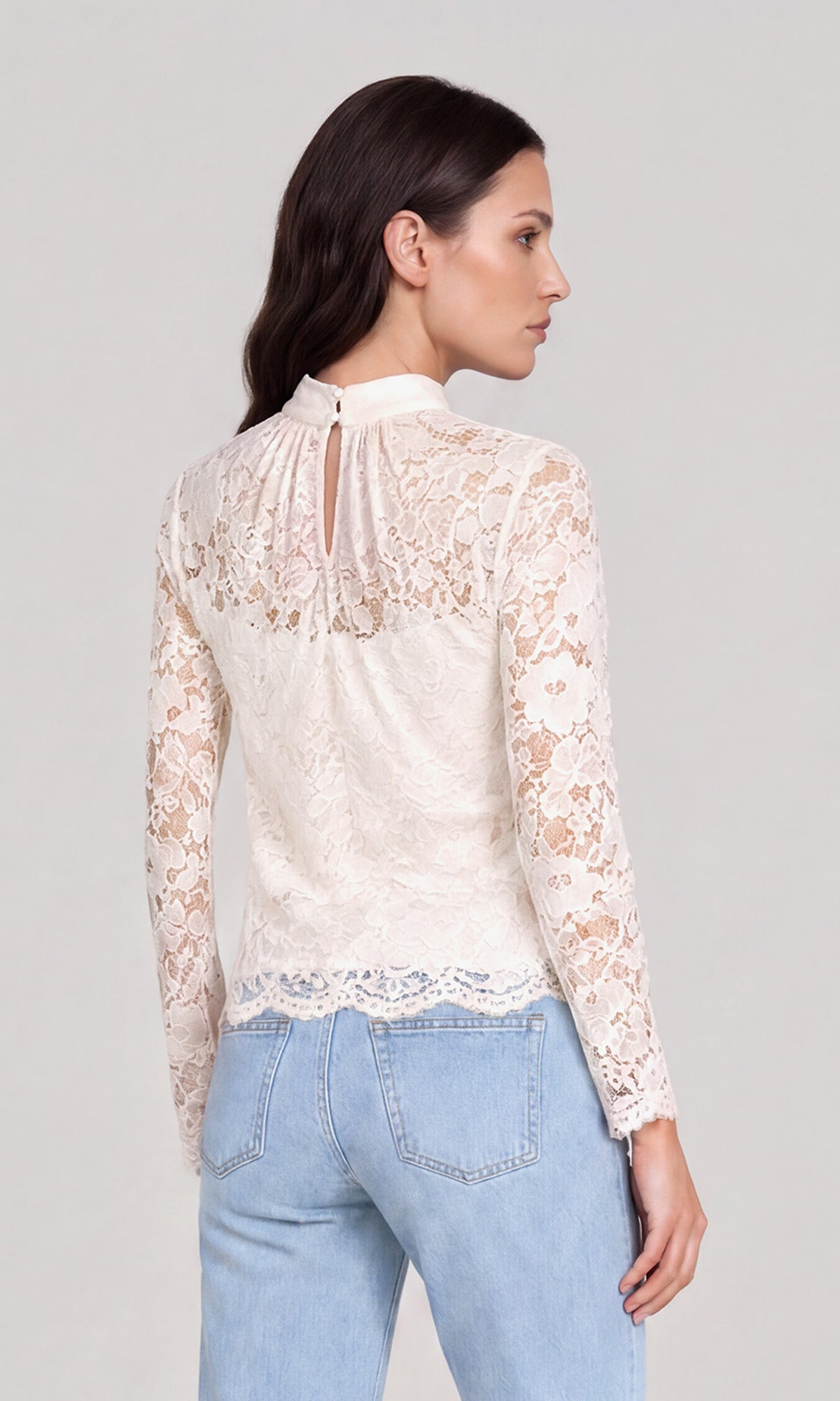 Back view of model wearing a cream lace mock-neck long-sleeve top paired with light blue jeans.