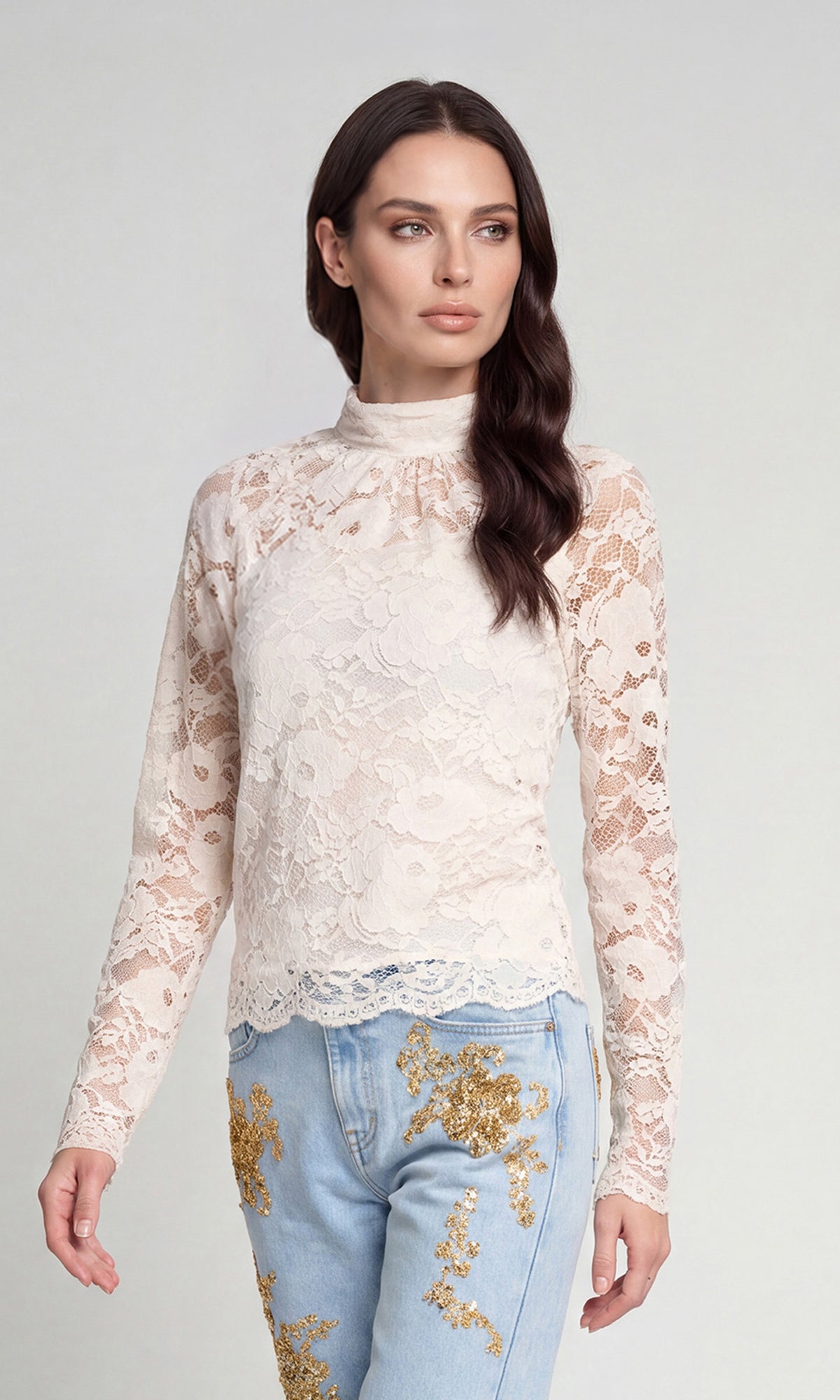 Side view of model wearing a cream lace mock-neck long-sleeve top paired with light blue jeans featuring gold sequin floral embroidery.