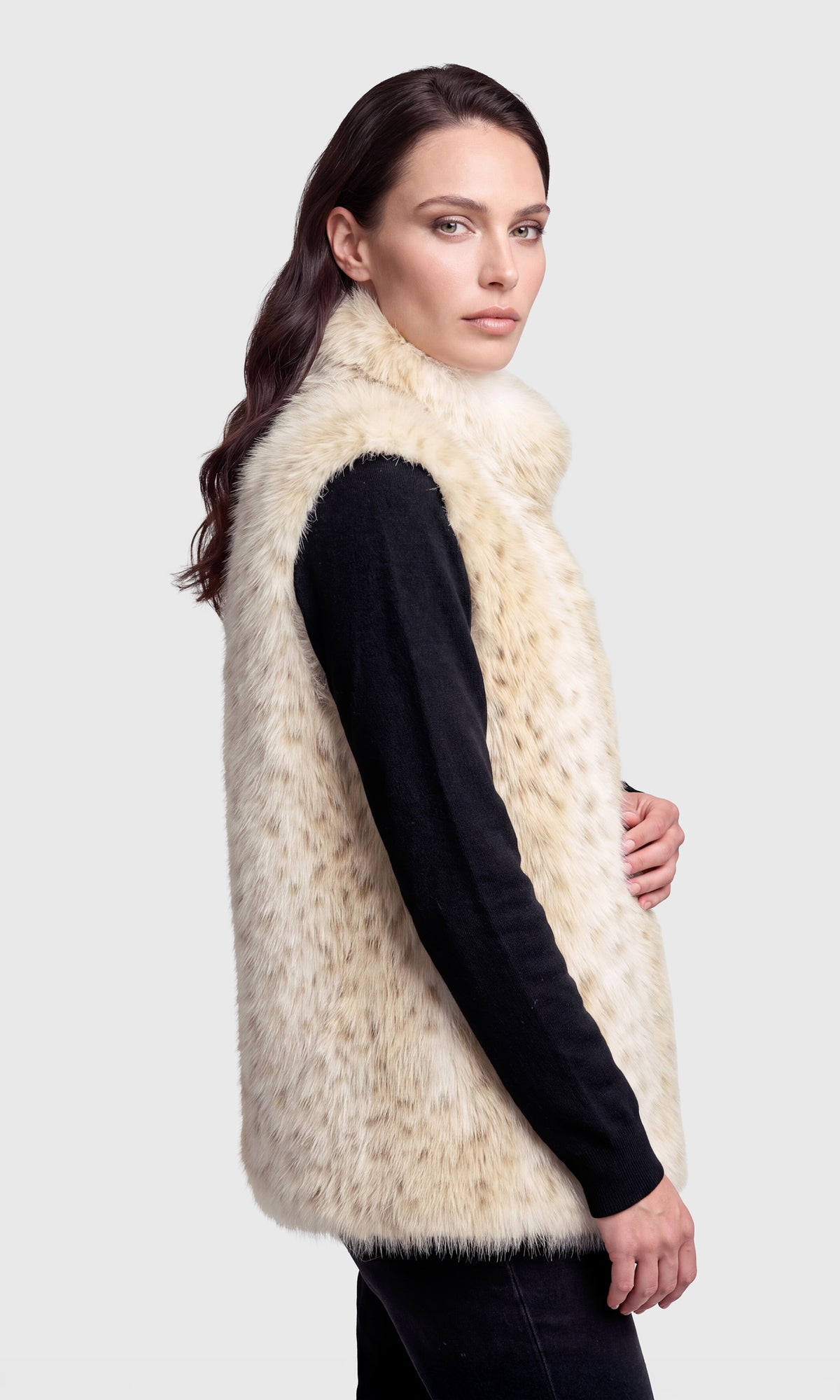 Side view of model wearing a sleeveless faux fur vest with a beige and cream leopard-like pattern, worn over a black long-sleeve top.