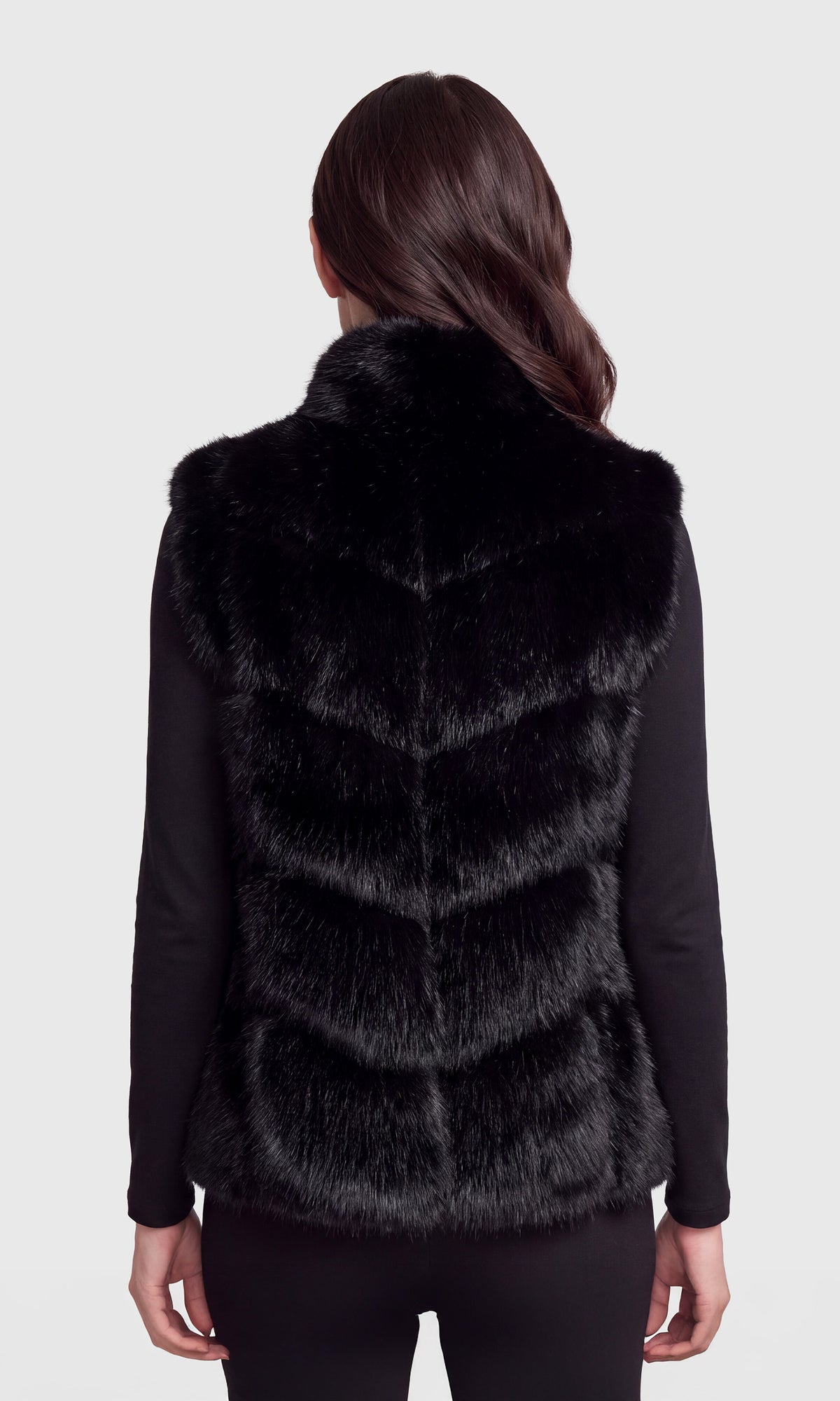 A woman viewed from the back wearing a black faux fur vest over a black long-sleeve top.