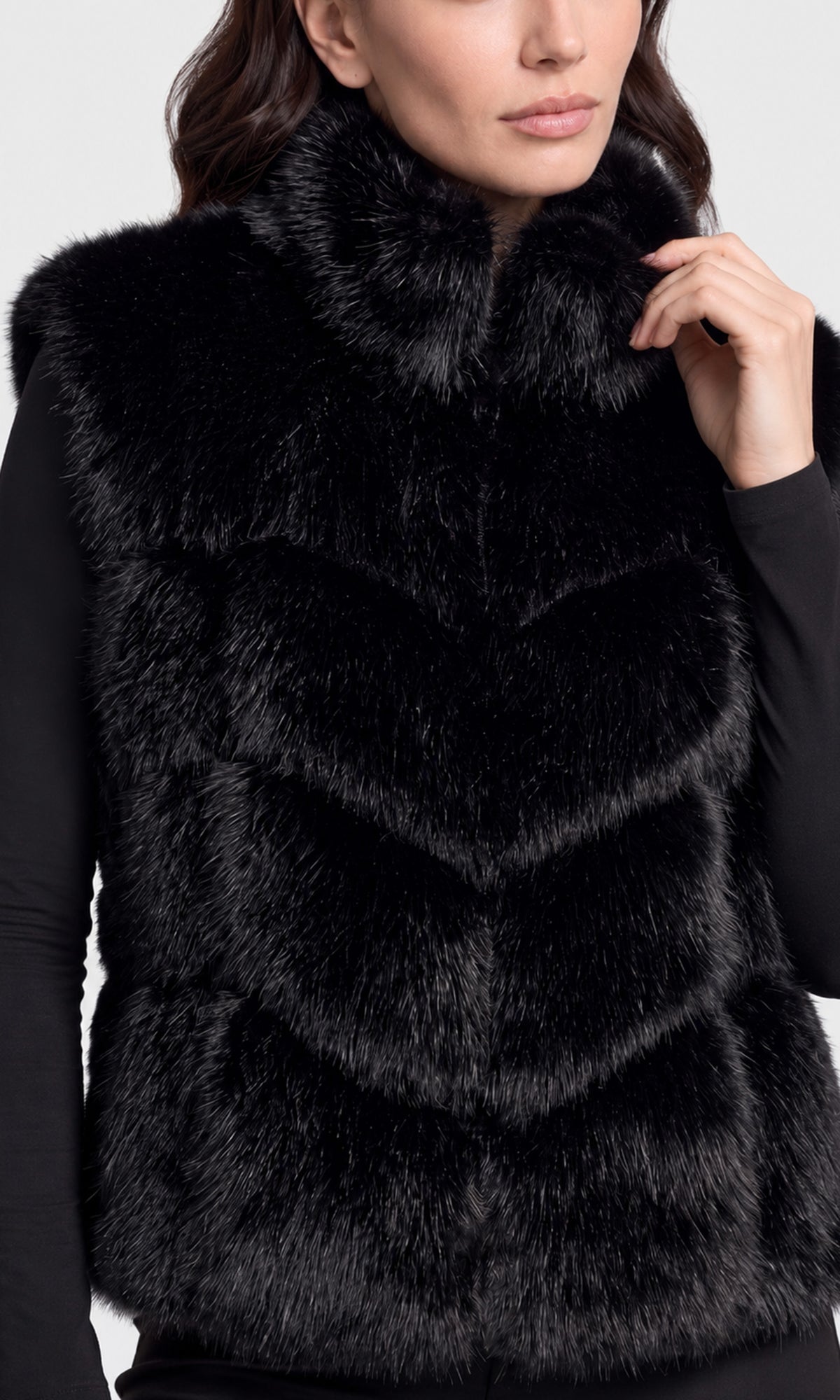 A close-up of a model wearing a black faux fur vest with a high collar over a black long-sleeve top.