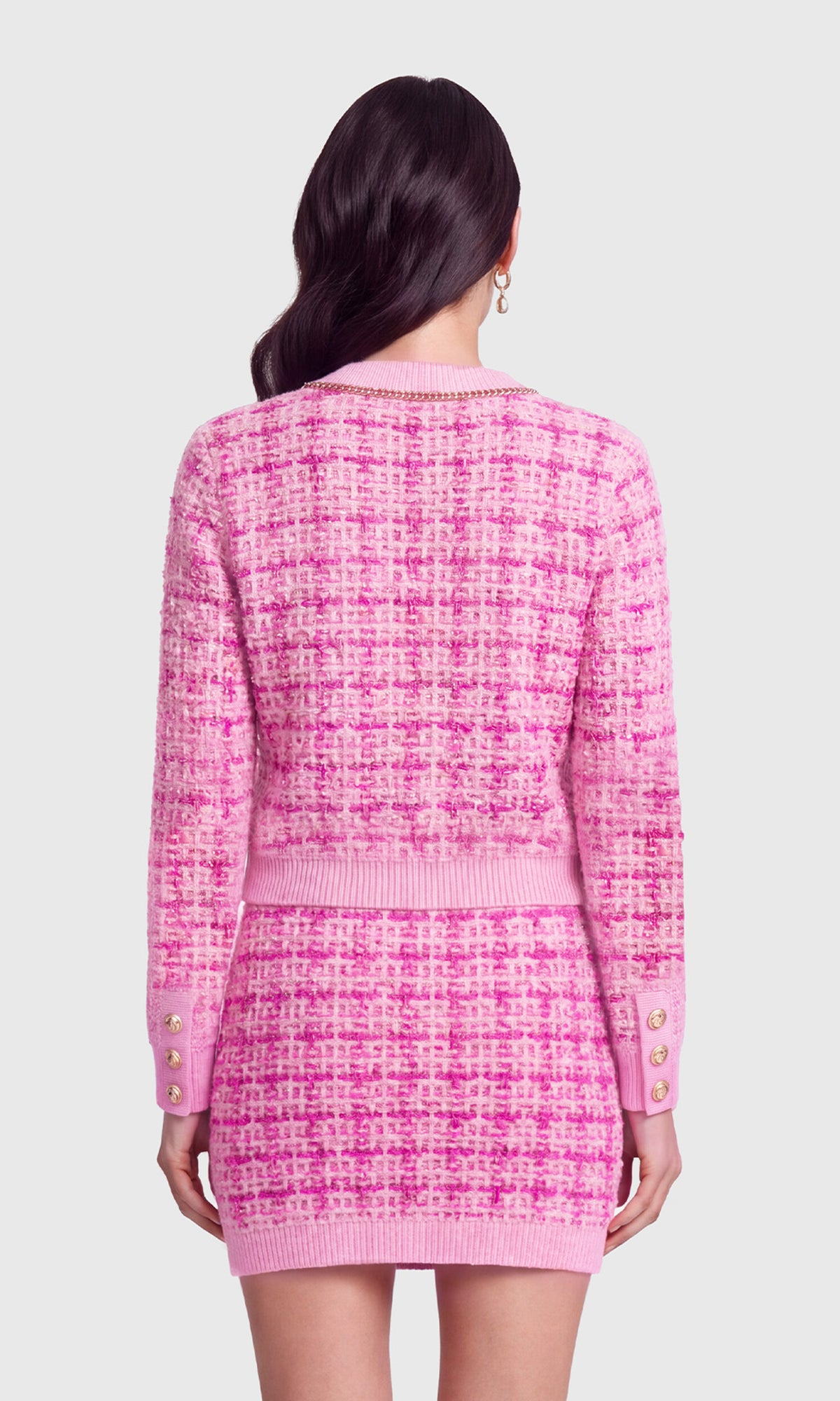 A woman shown from the back wearing a pink tweed long-sleeve cardigan and matching mini skirt set with gold buttons.