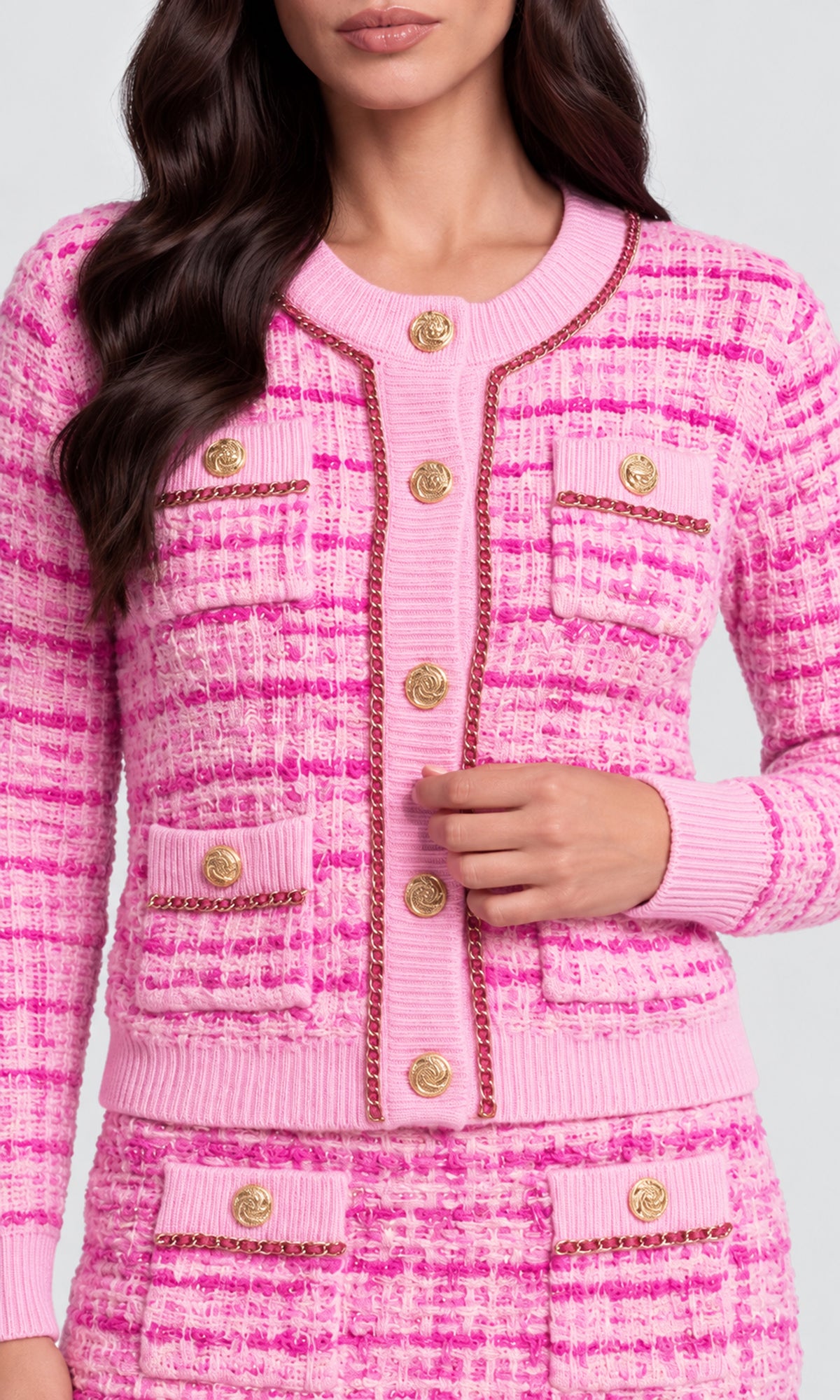A close-up of a model wearing a pink tweed cardigan with gold buttons and gold trim, paired with a matching pink tweed skirt. 