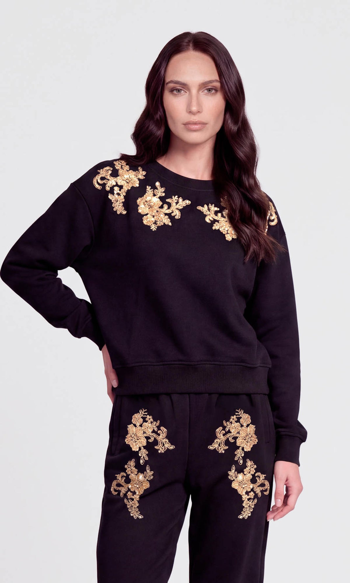 A woman modeling a black crewneck sweatshirt and matching sweatpants set, both decorated with ornate gold floral embroidery on the shoulders and upper thighs.