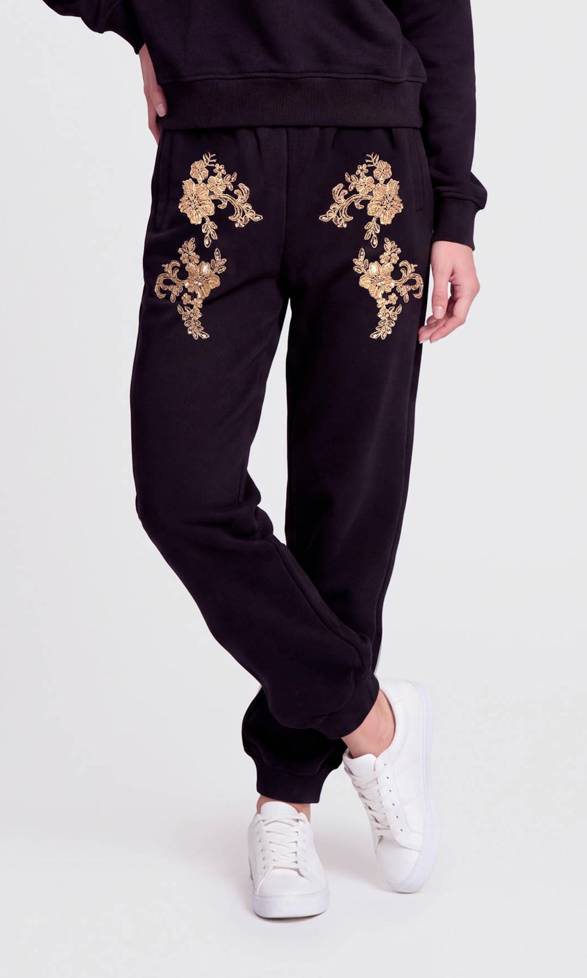 Model wearing black sweatpants with gold floral embroidery on the upper thighs, paired with white sneakers.