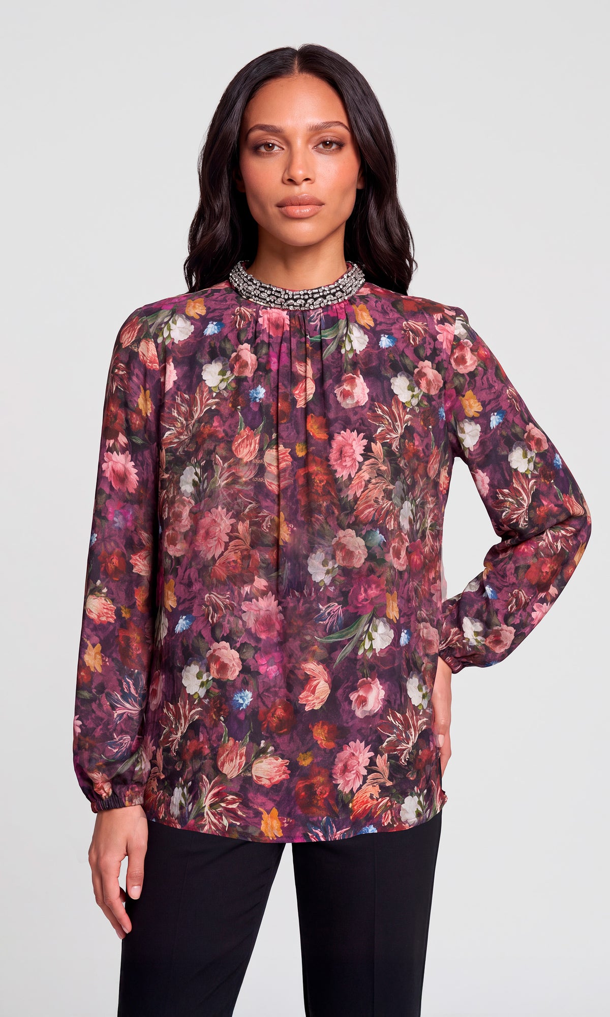 Model wearing a long-sleeve floral blouse with a high gathered neckline and a beaded collar trim, paired with black pants.