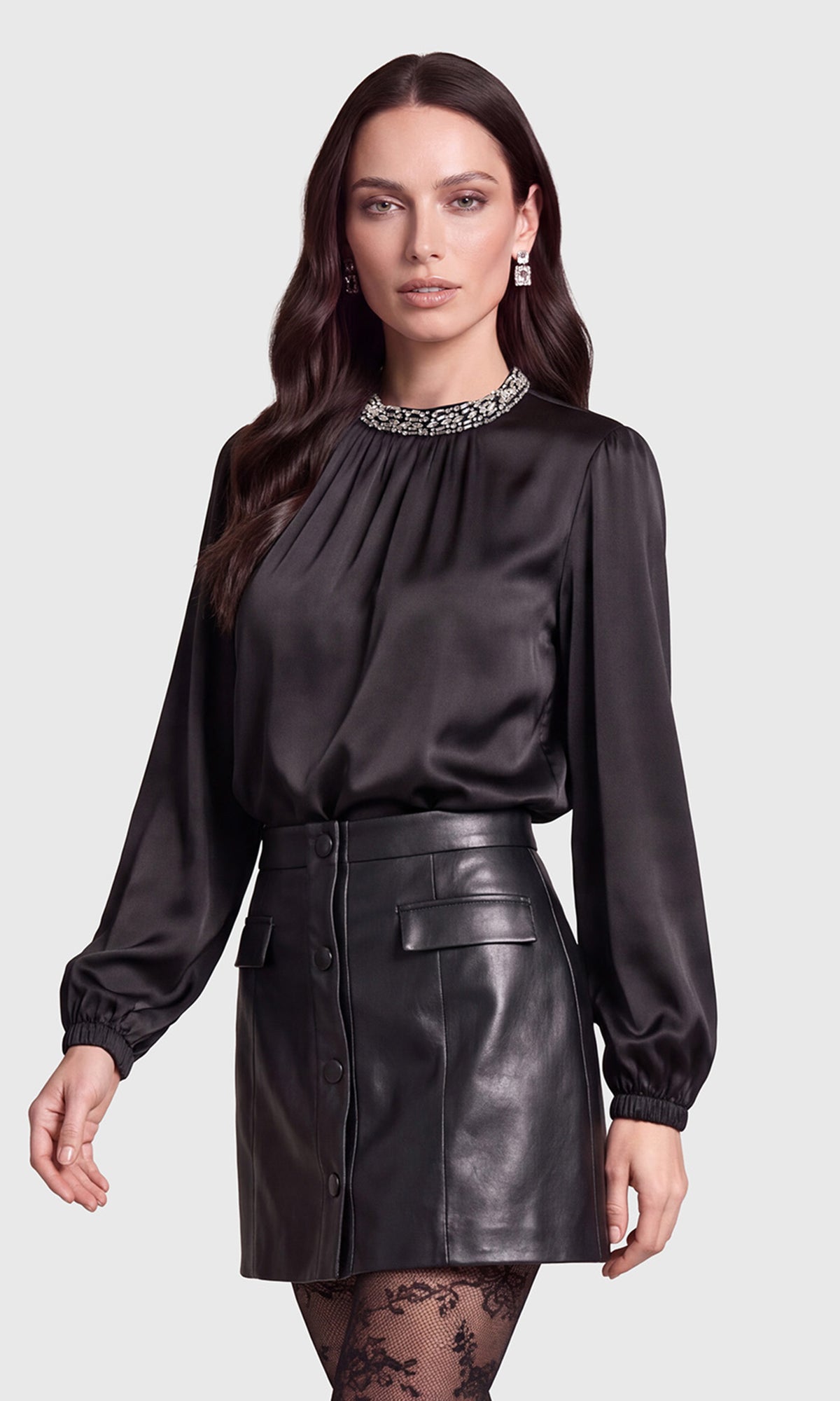 Side view of model wearing a black long-sleeve satin blouse with a jeweled high neckline, paired with a black faux-leather button-front mini skirt and black lace tights.