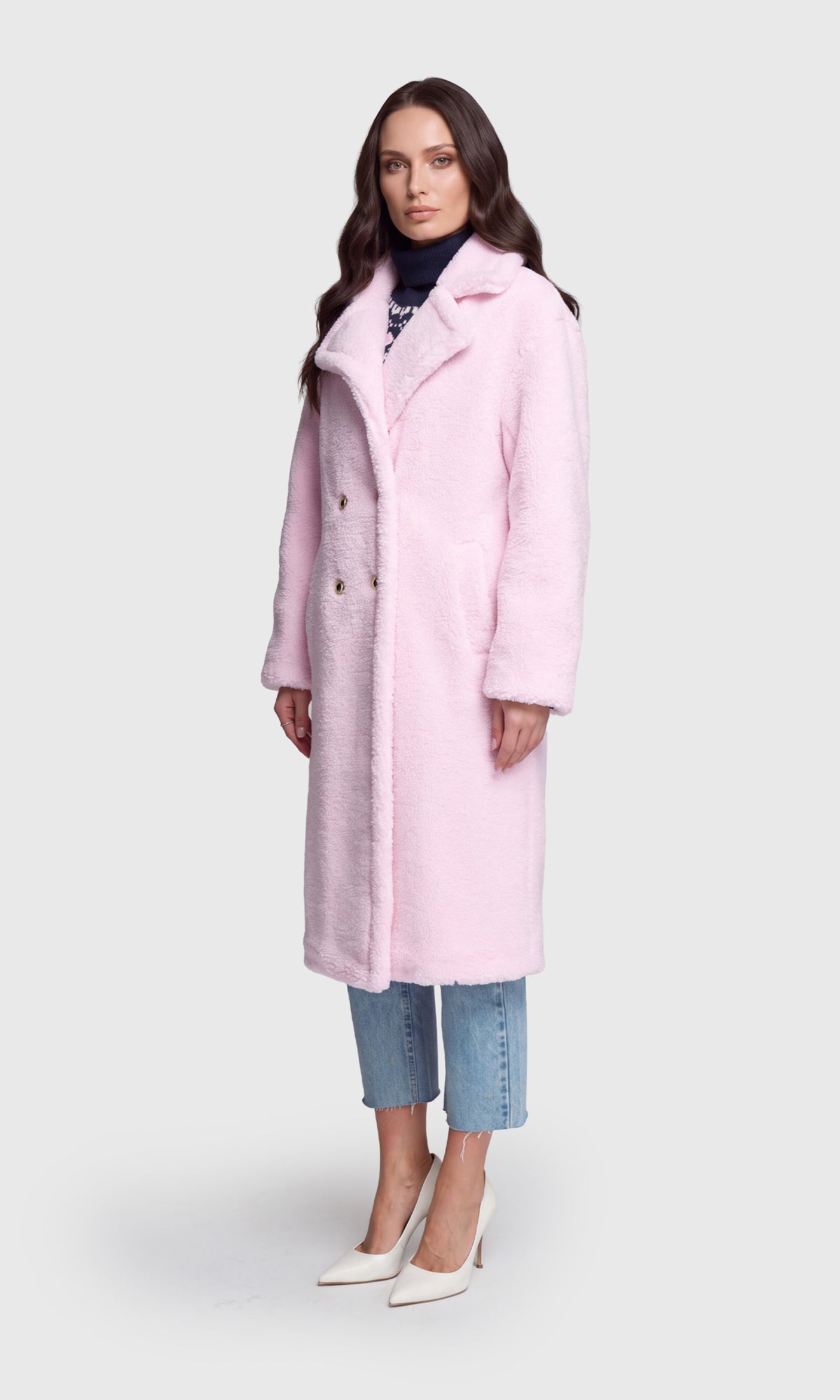 A woman modeling a light pink, mid-length fuzzy coat over a dark turtleneck, cropped blue jeans, and white pointed-toe heels.
