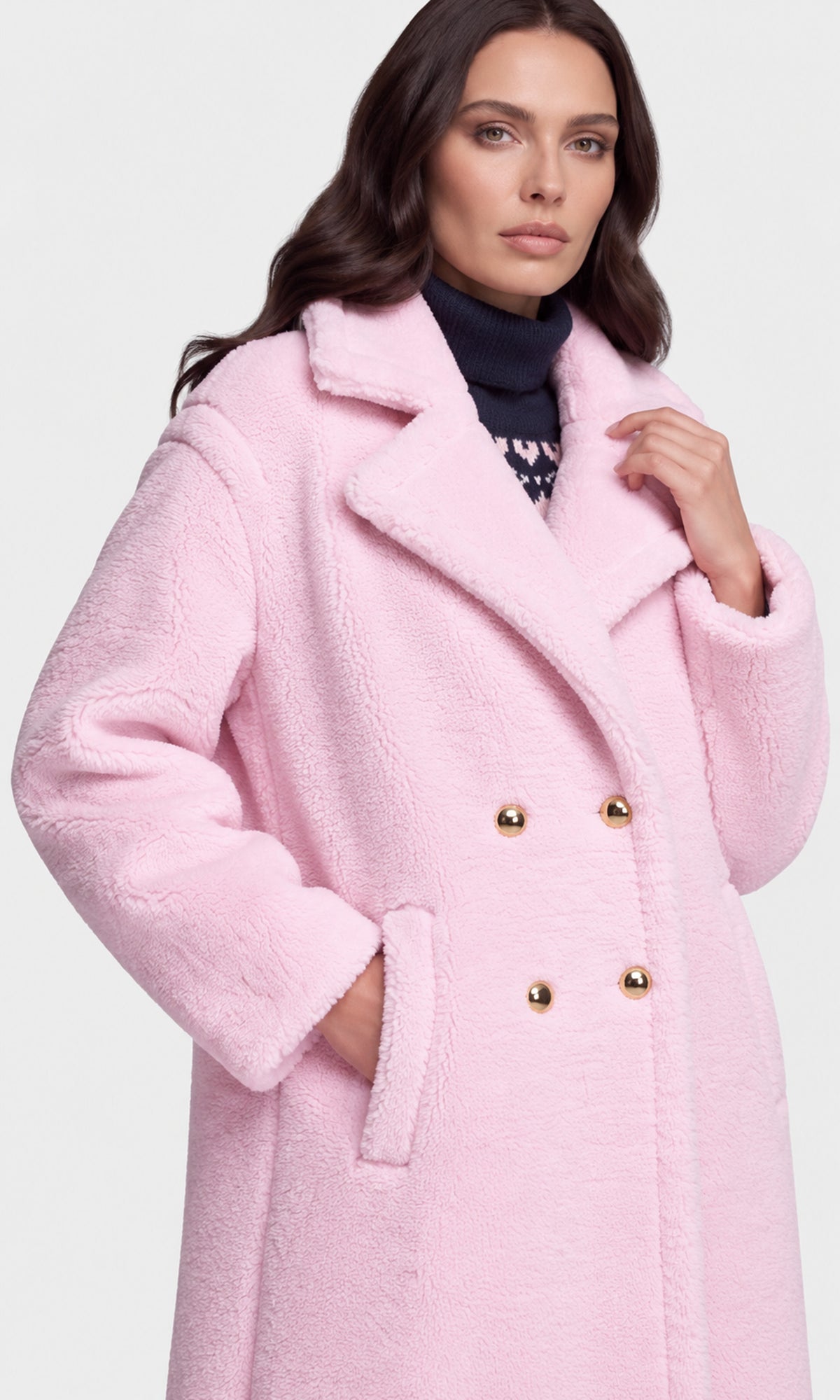 A woman wearing a light pink teddy coat with gold buttons over a dark navy turtleneck.