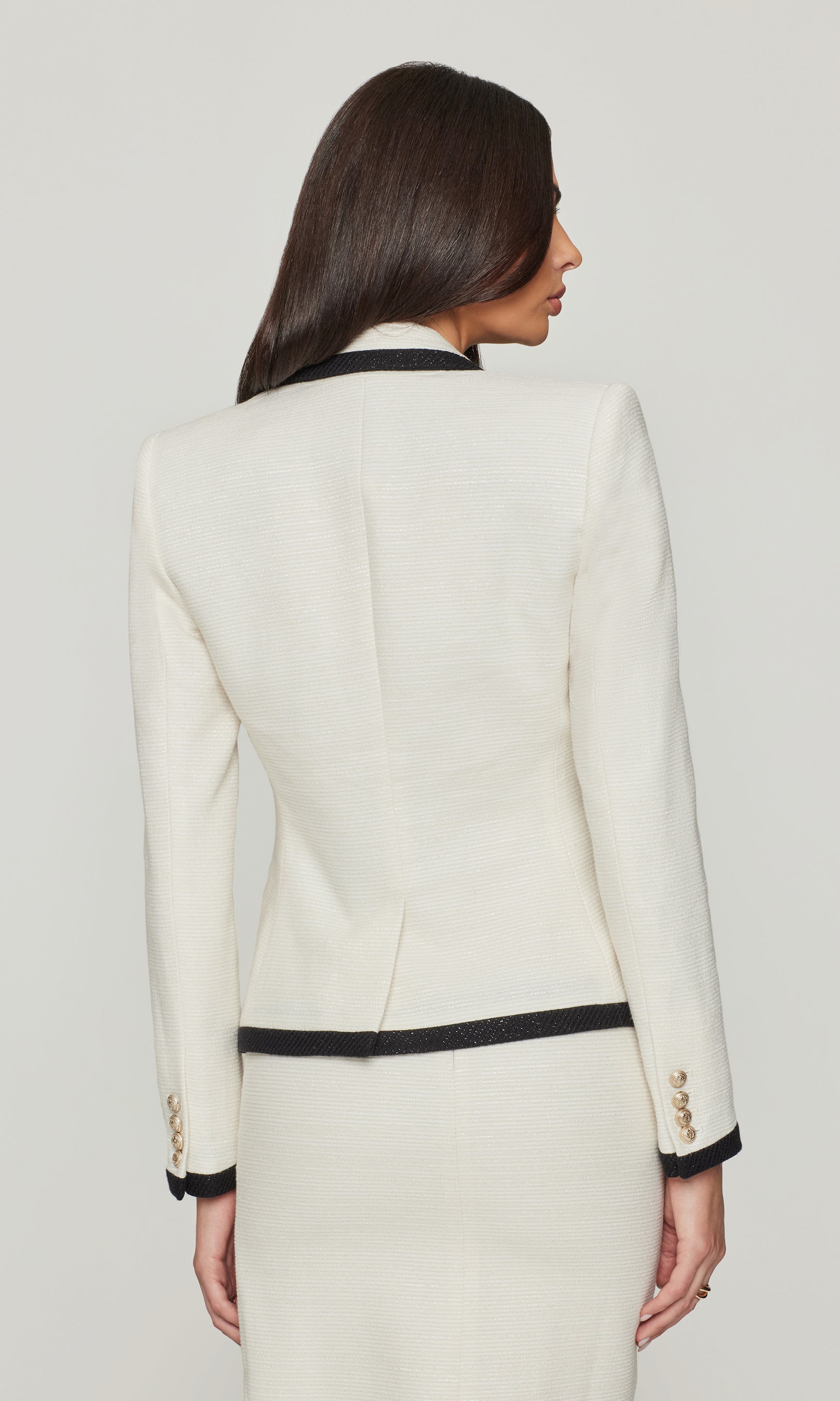Back view of woman wearing a cream blazer with black trim on a plain background