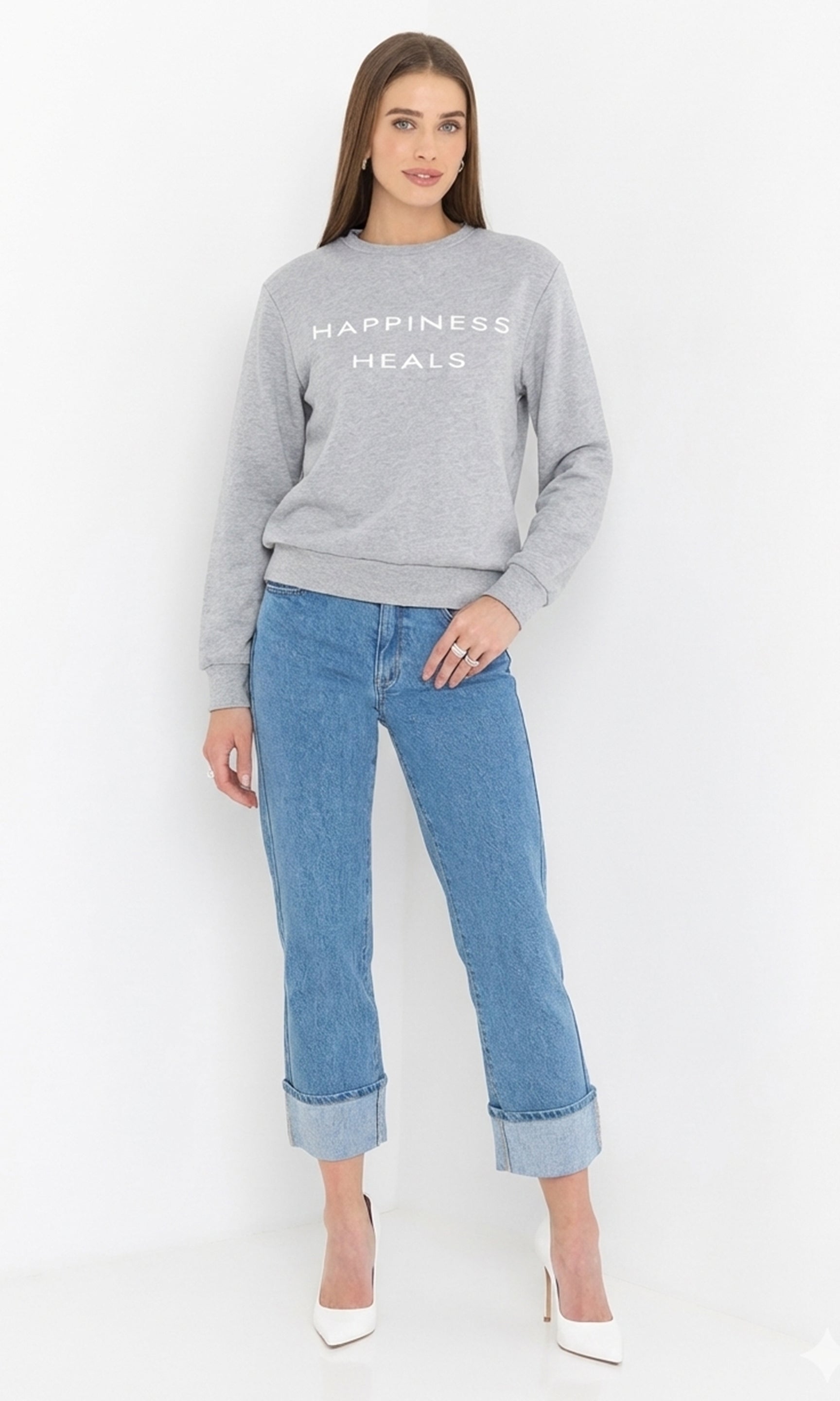 Woman wearing a gray sweatshirt with 'Happiness Heals' text and blue jeans on a white background