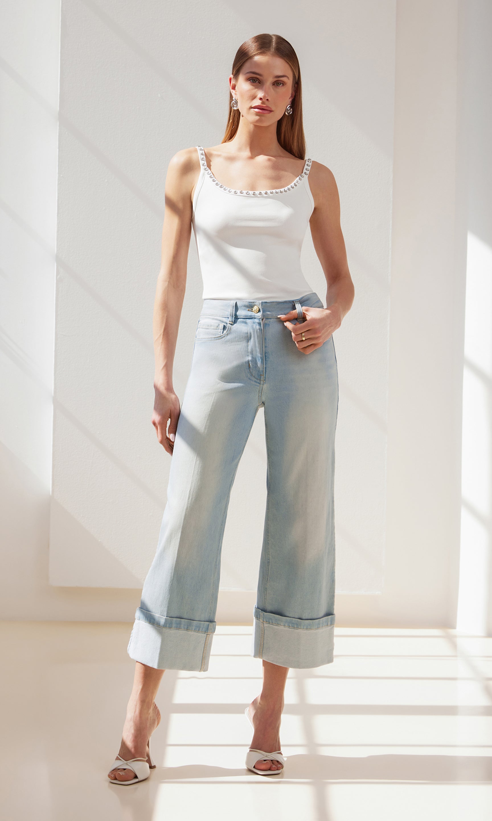 full body image of model posing in front of a window wearing the Iris tank in white, paired with blue jeans, white heels and silver earrings