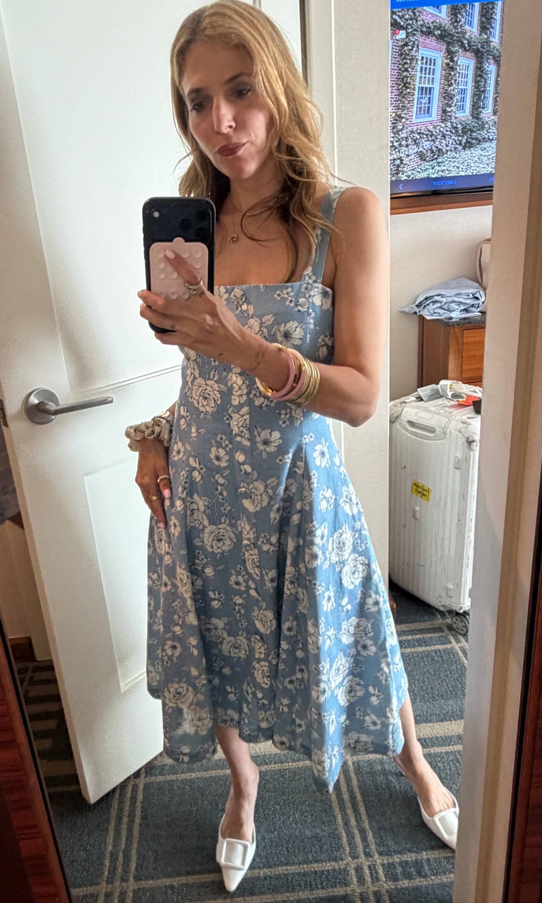 A woman takes a mirror selfie in a hotel room wearing a light blue midi dress with a white floral pattern, white pointed-toe flats, and stacked bracelets.