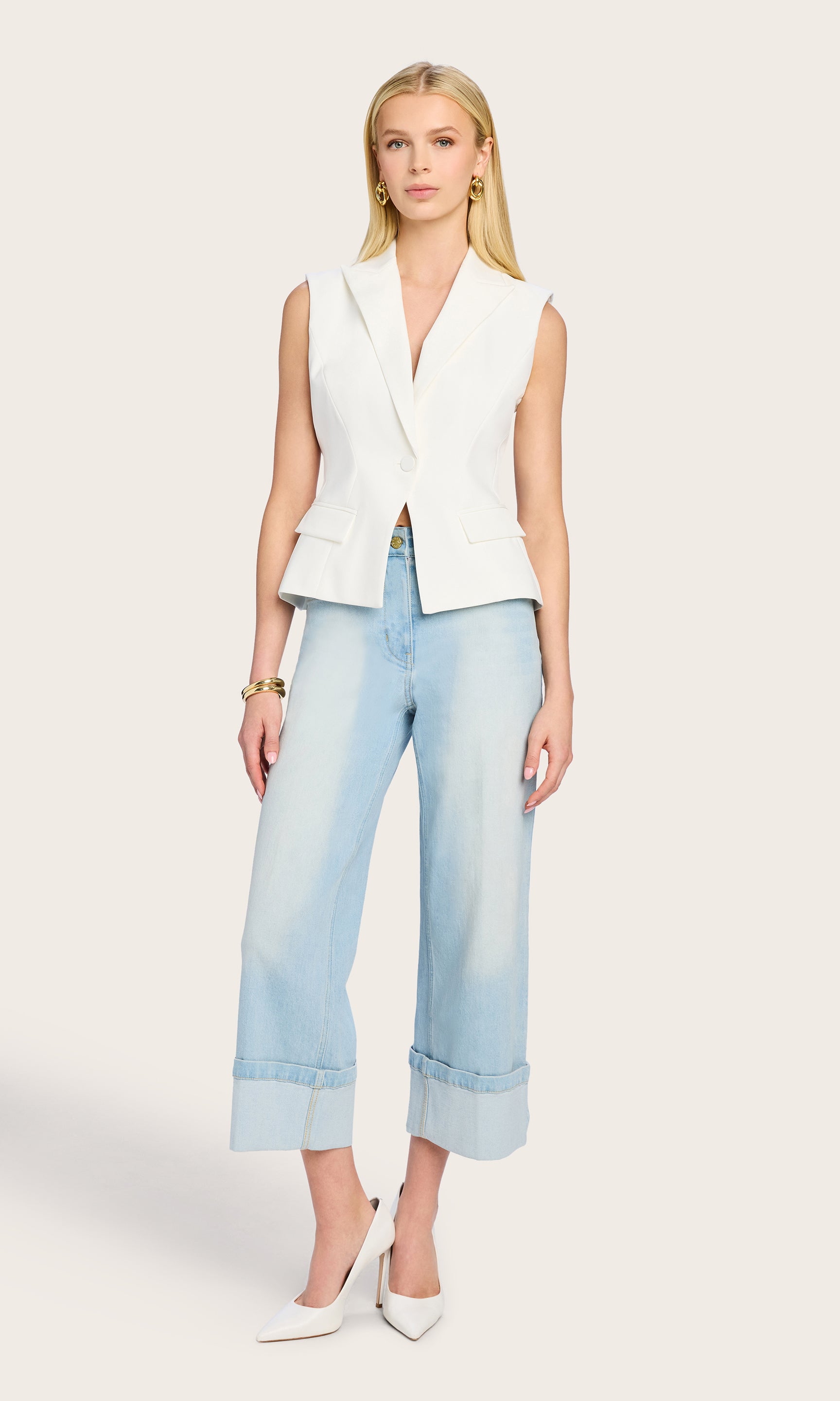 The model poses in full body view wearing the Jenna Sleeveless Blazer styled with light-wash jeans and heels, capturing the elongated fit and sleek lines of the piece.
