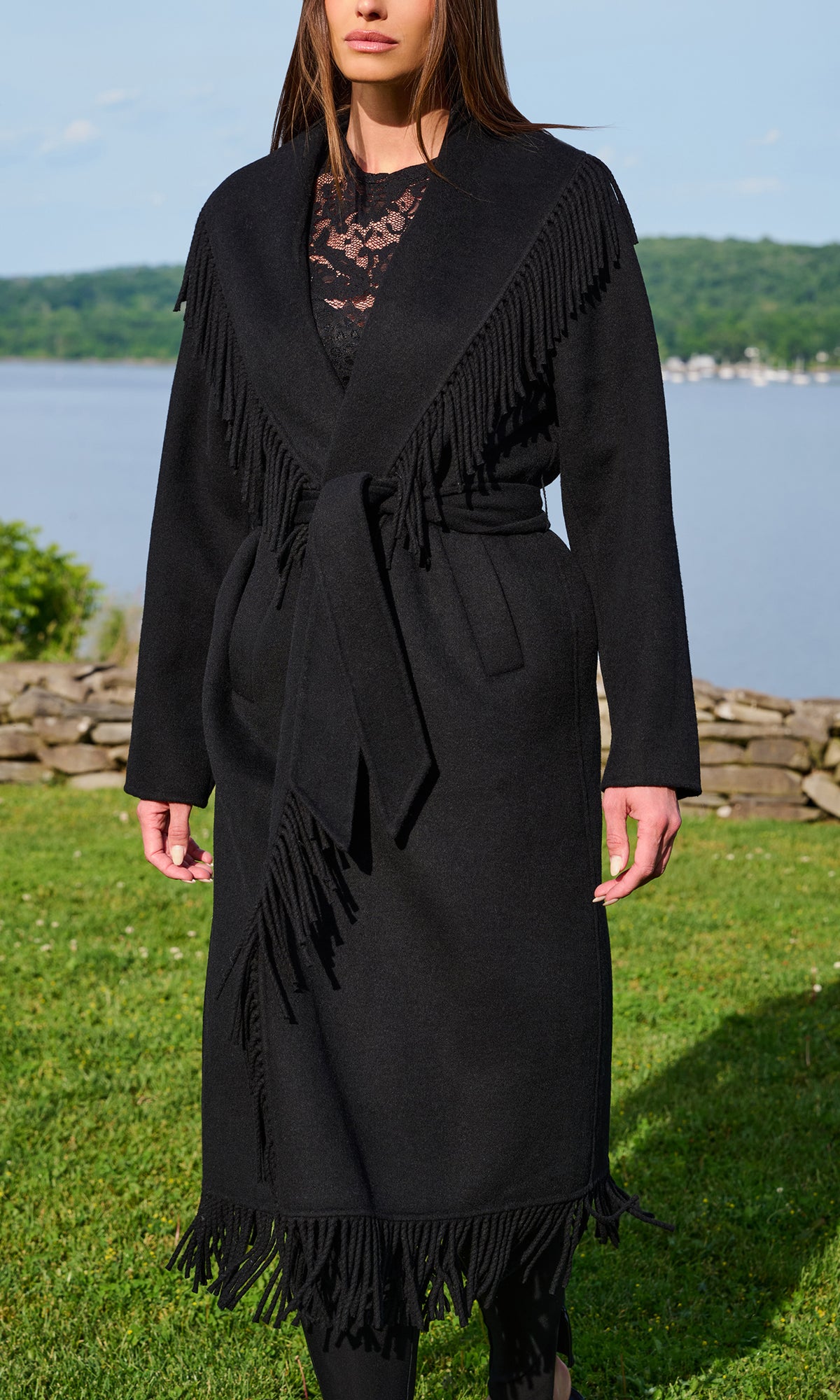 A woman wearing a long black wrap-style coat with long fringe trim and a self-tie belt, photographed outdoors on grass with water and trees in the background.