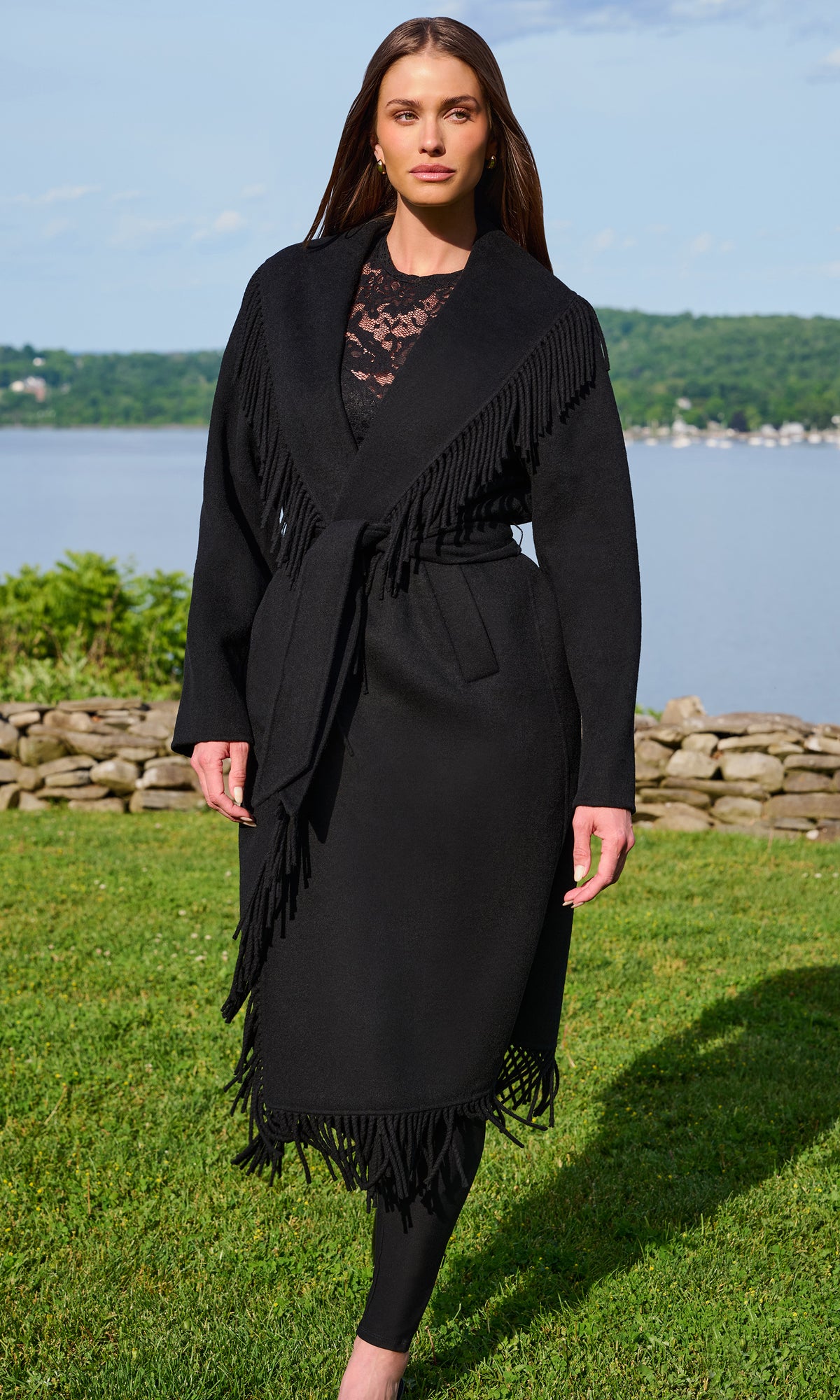 A woman stands outdoors on a grassy lawn with a lake and trees in the background, wearing a long black fringed wrap coat over a black lace top and black leggings.