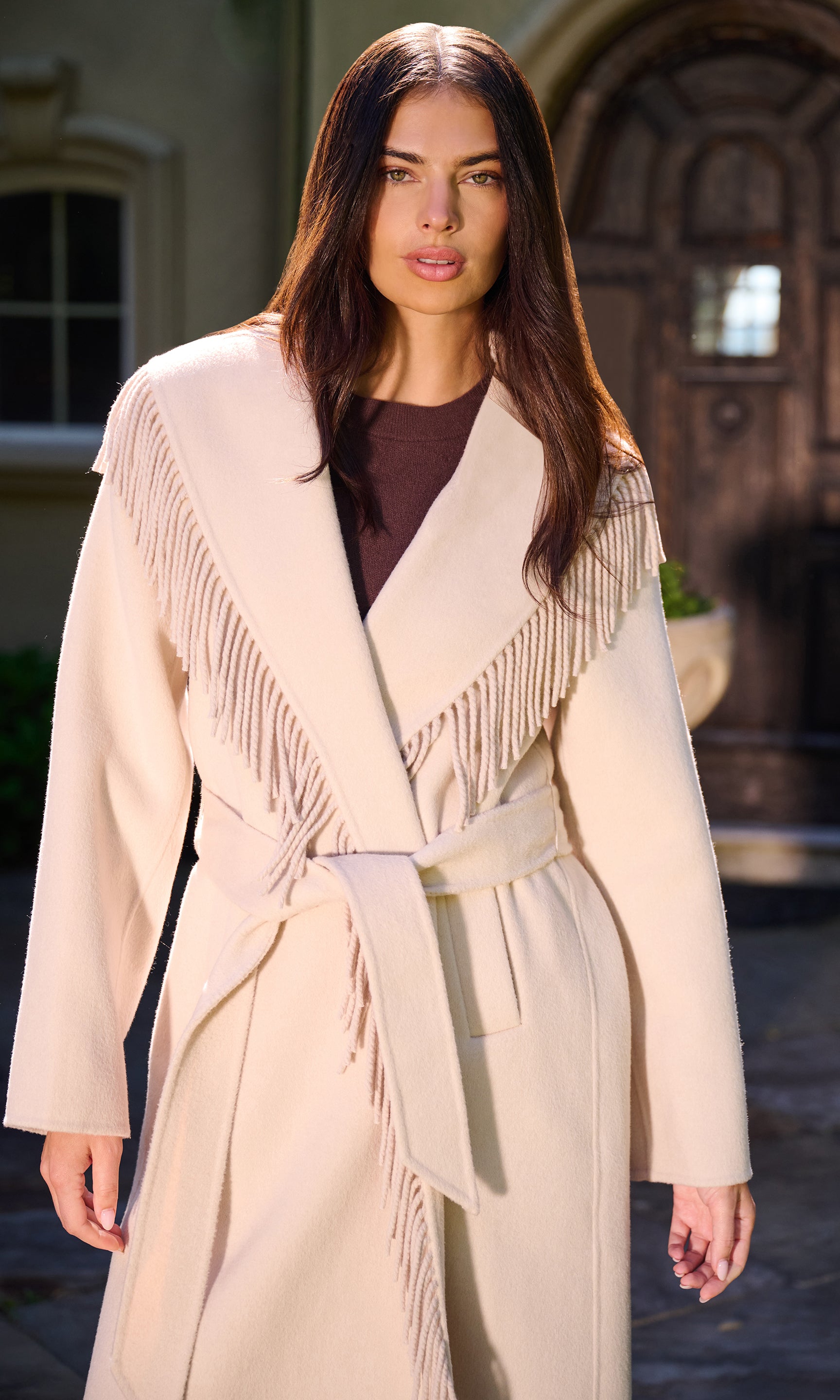 A woman wearing a long cream-colored wrap coat with fringe detailing and a matching tie belt, standing in front of a stone building entrance.
