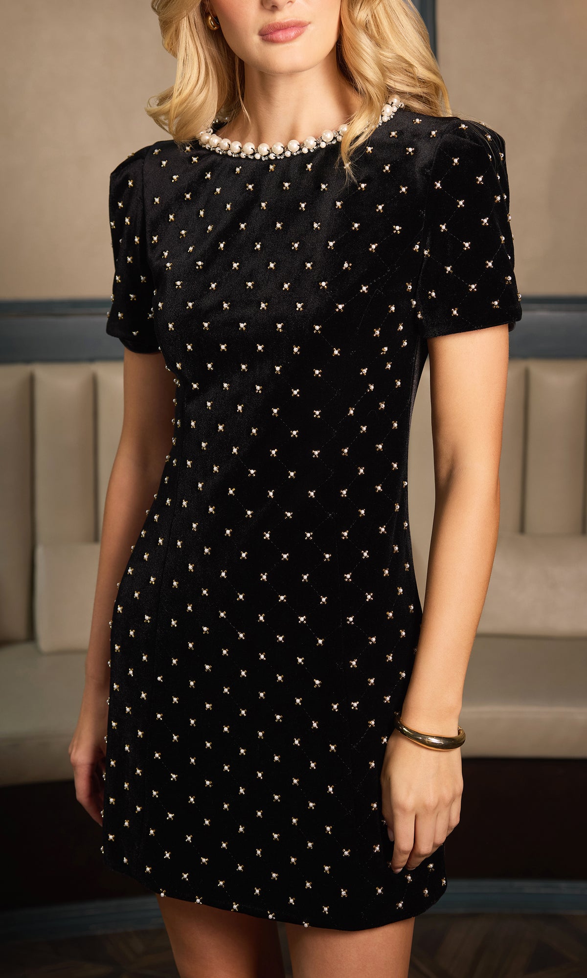 A woman wearing a short-sleeve black velvet dress with a gold geometric star pattern and a pearl-trimmed neckline, photographed in an indoor setting.