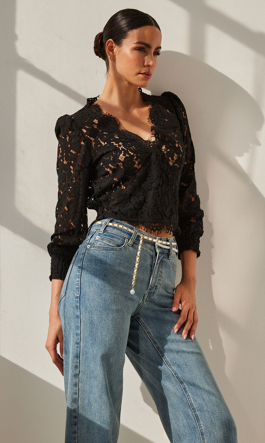 Woman leaning against the wall while wearing a v-neck, long-sleeve black lace blouse and jeans.