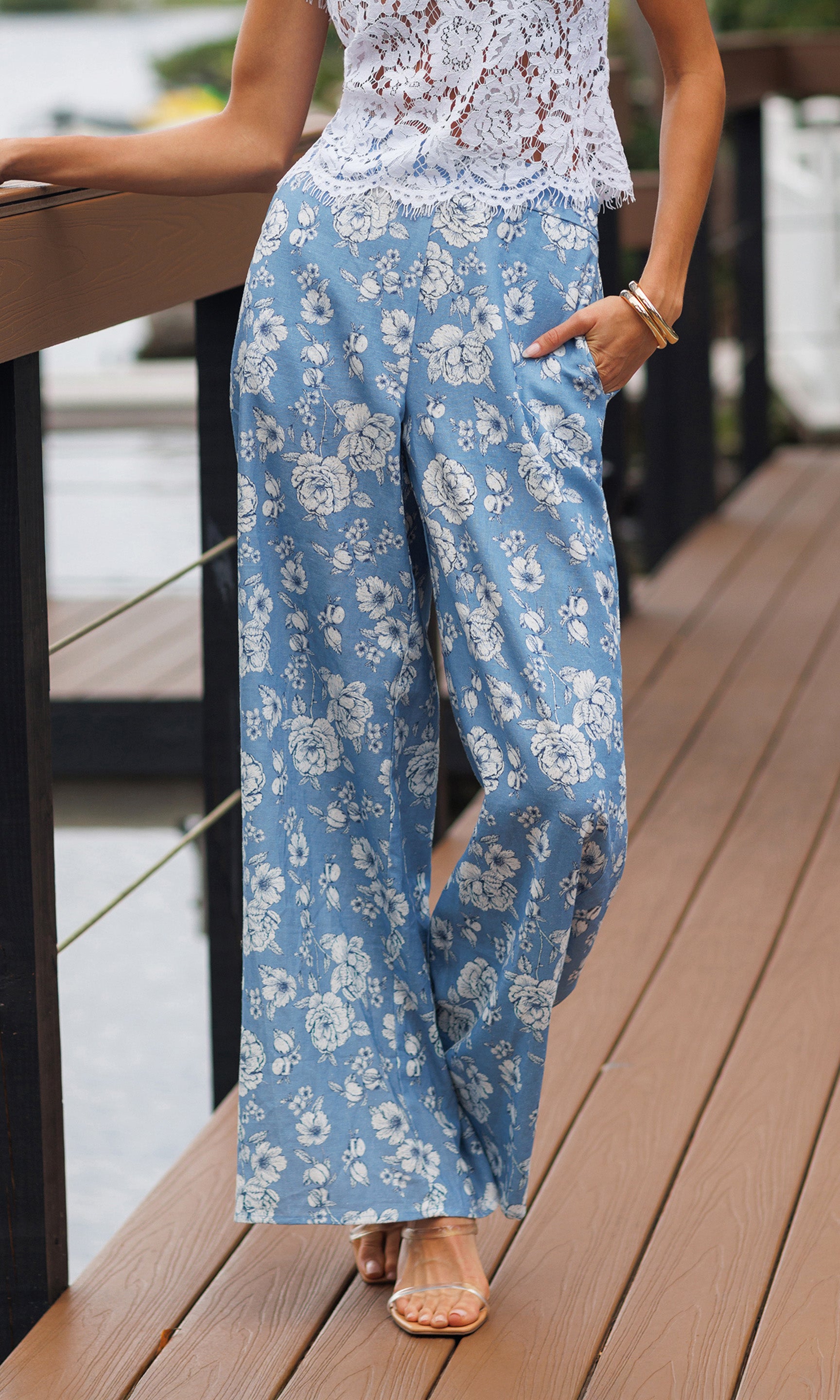 Person wearing a white lace top and blue floral pants on a wooden deck.