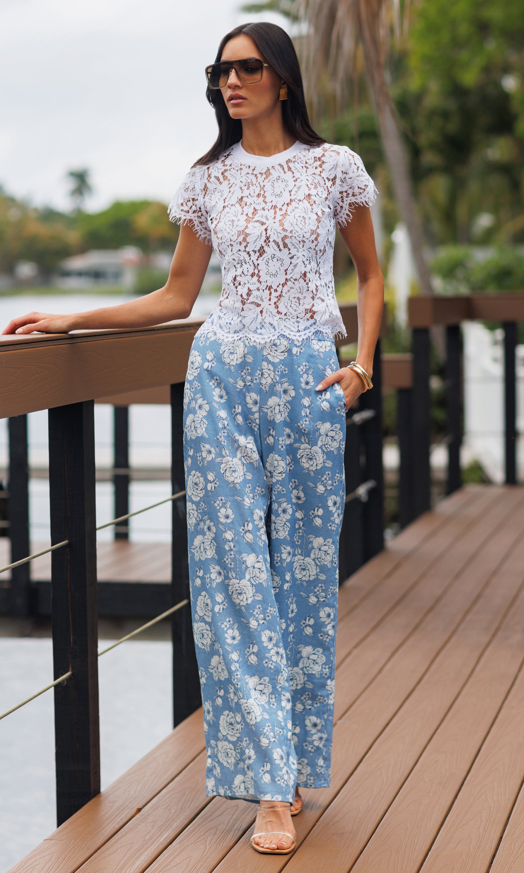 Woman in a white lace top and blue floral pants standing on a wooden deck by a waterfront.