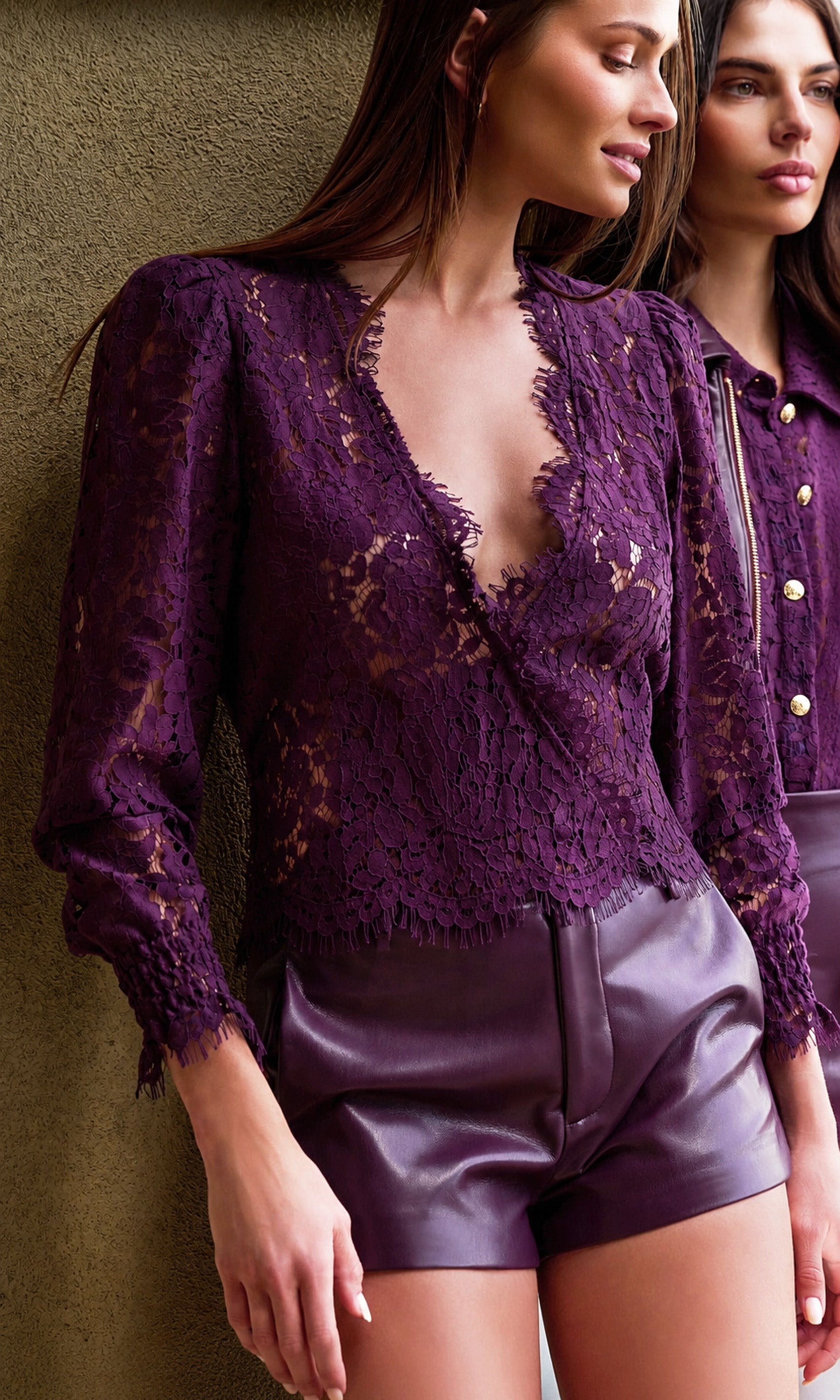 A woman wearing a deep purple lace long-sleeve wrap-style top and matching purple faux-leather shorts, standing against a textured wall.