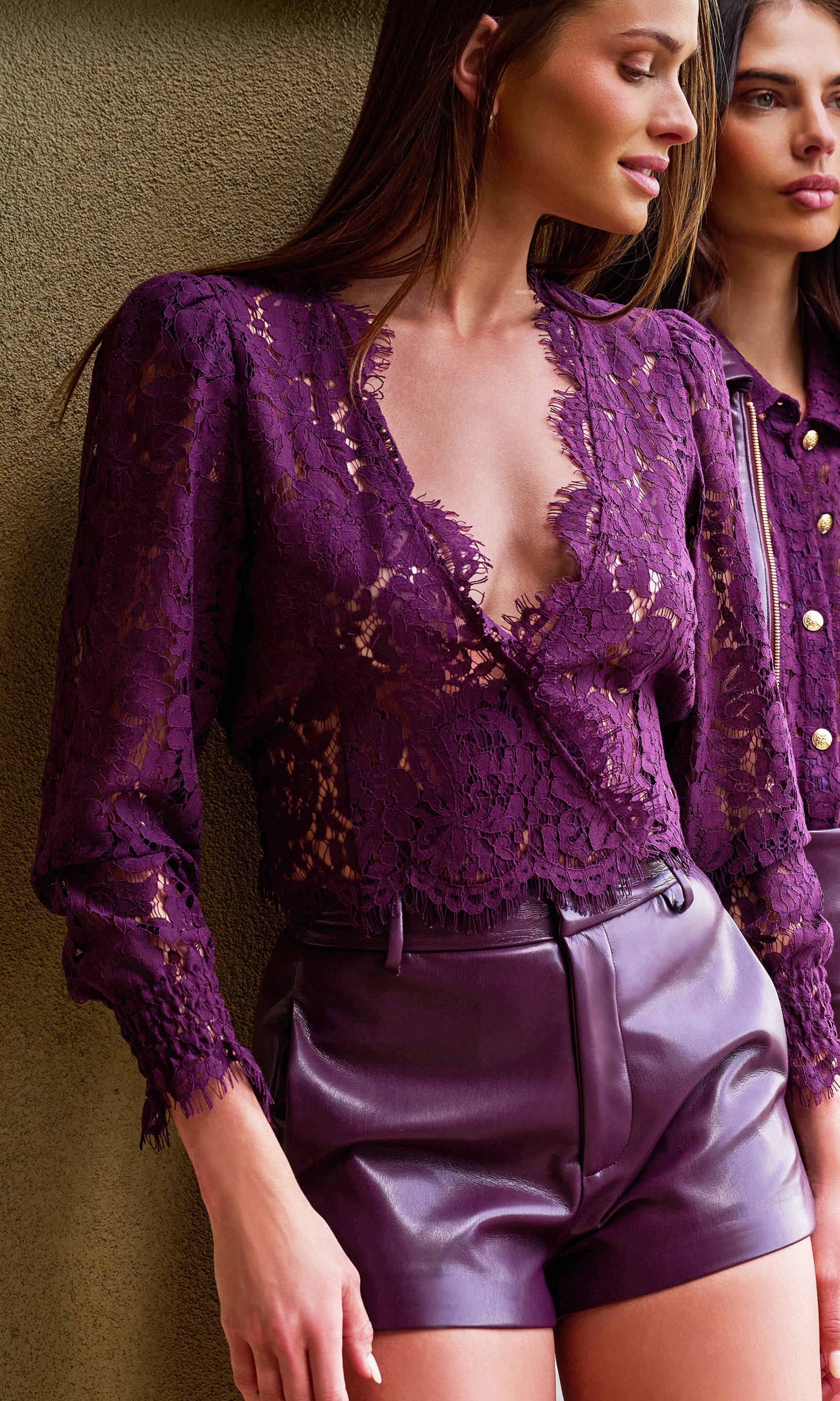 A woman wearing a deep purple lace long-sleeve top with a deep V-neck and matching purple faux-leather shorts, standing against a textured beige wall.