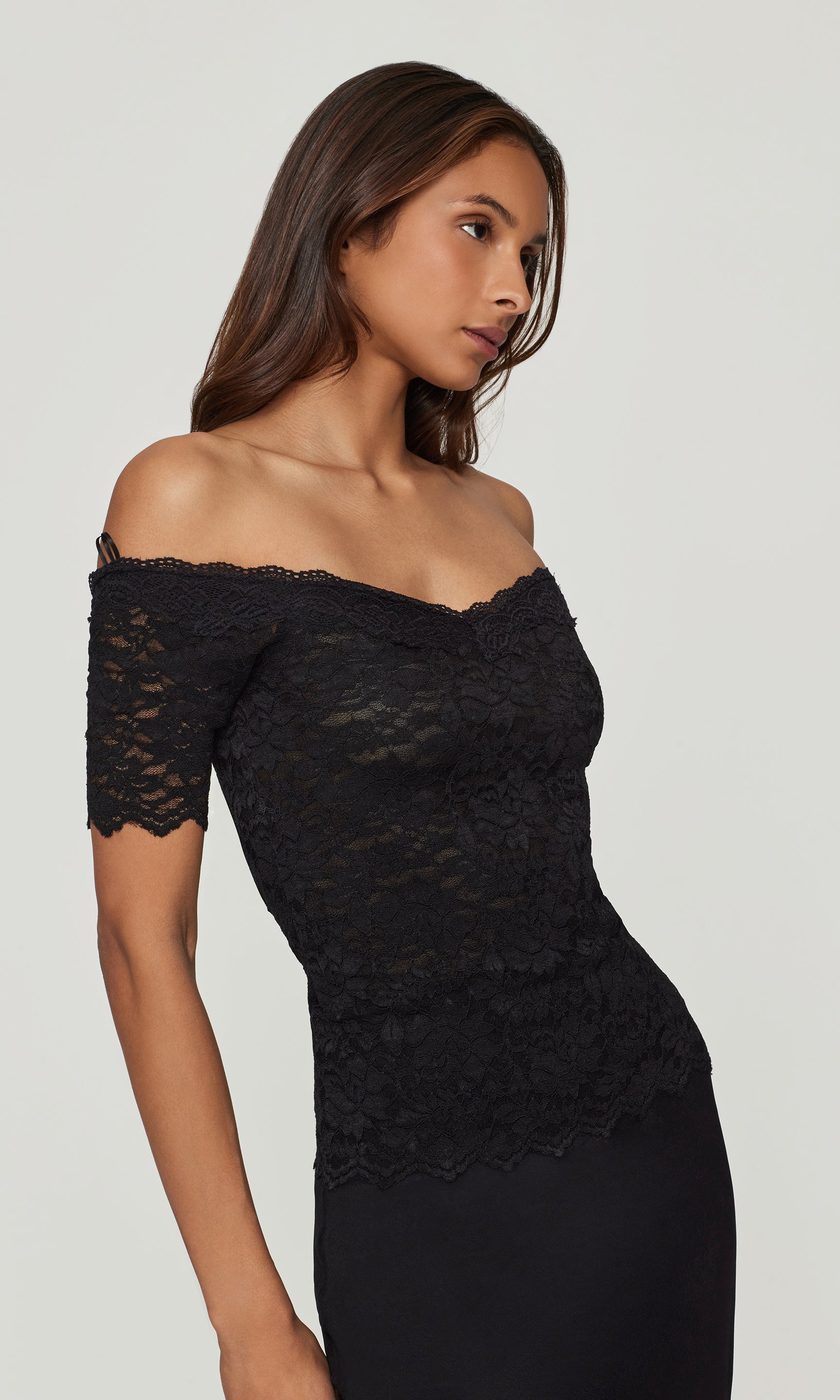Side view of woman wearing a black lace off-shoulder top and black skirt against a plain background